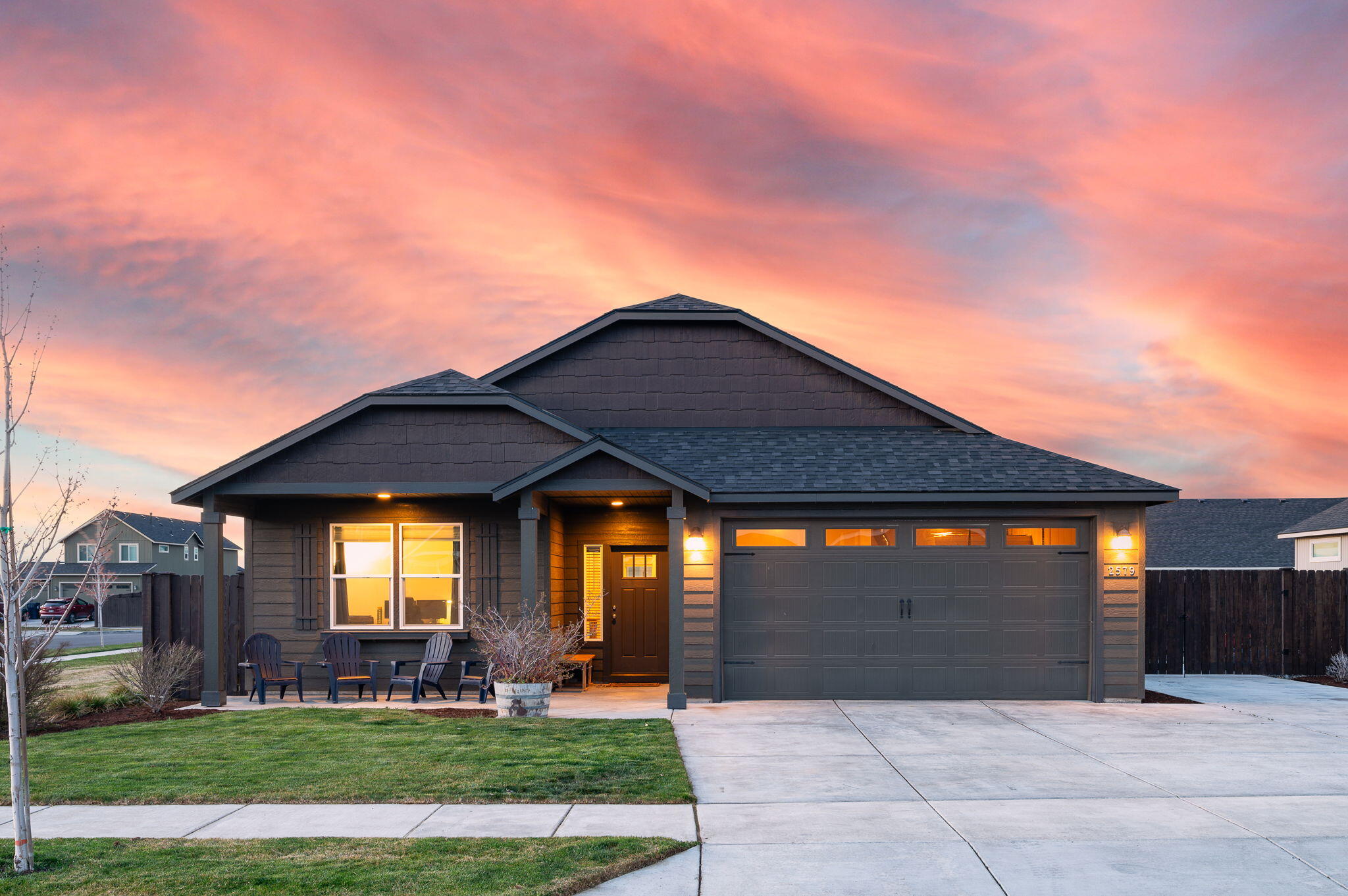 Dark gray single-story home with a two-car garage and warmly lit entry beneath a pink dusk sky.