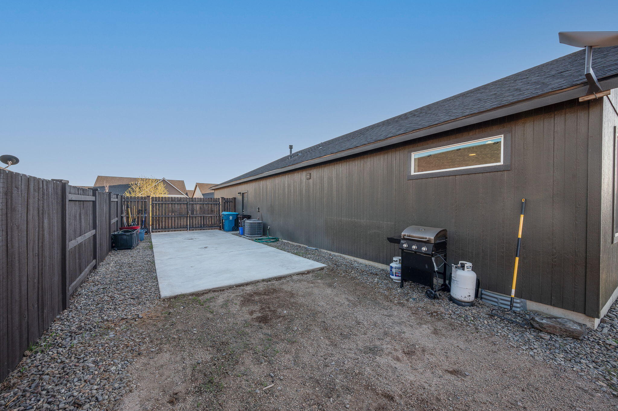 Fenced side yard with a concrete patio slab and propane grill beside a dark-sided home.