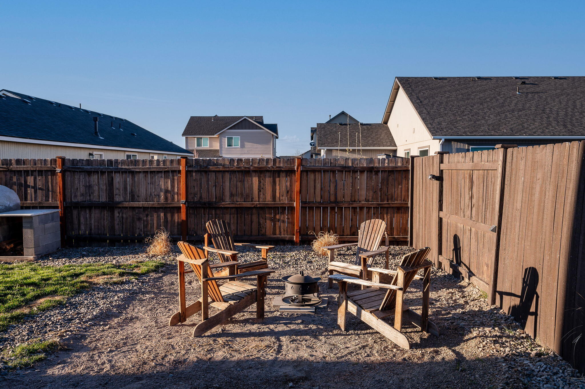 Fenced backyard seating area with wooden chairs around a fire pit on gravel.