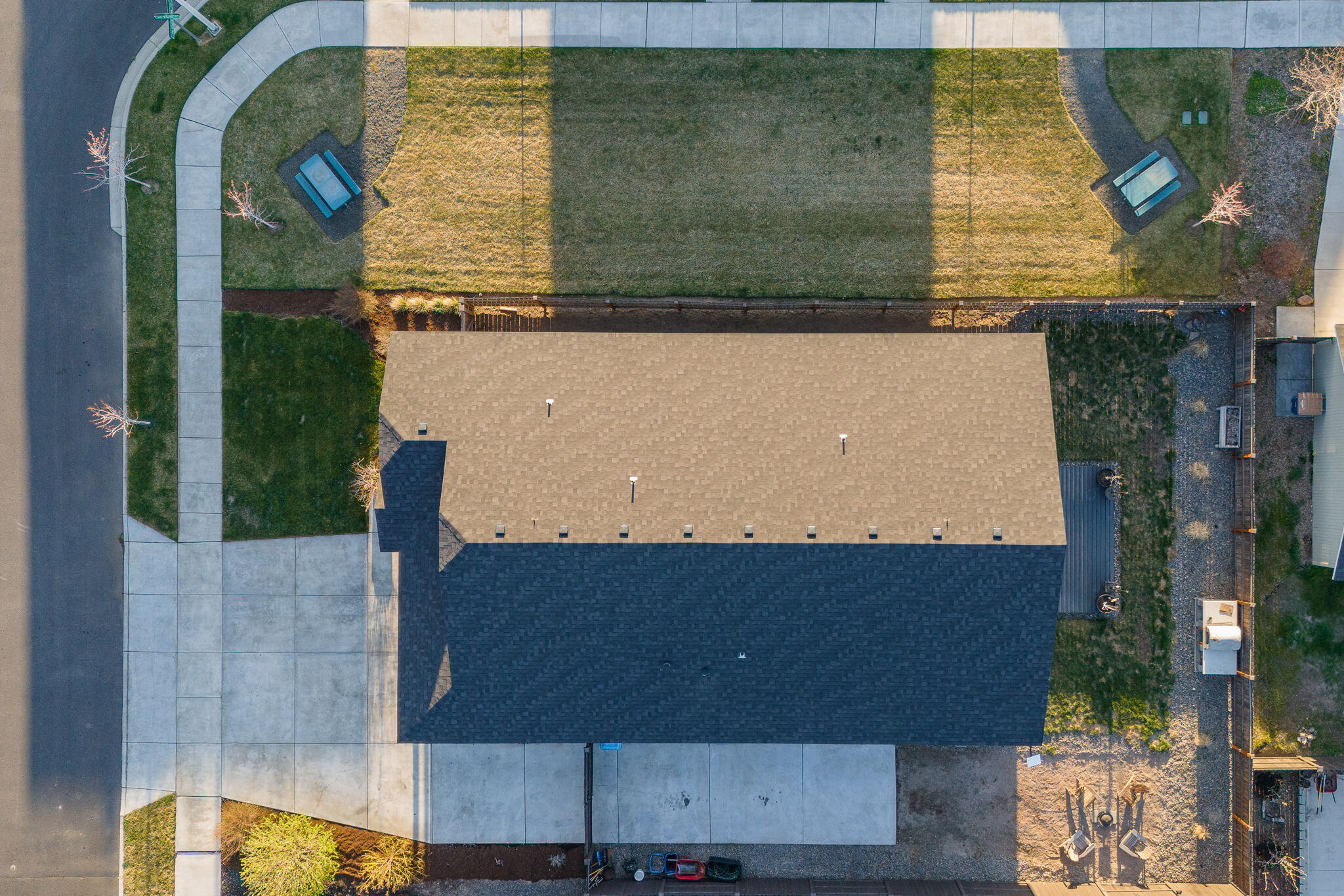 Aerial view of a home with a broad shingled roof and fenced backyard with benches.