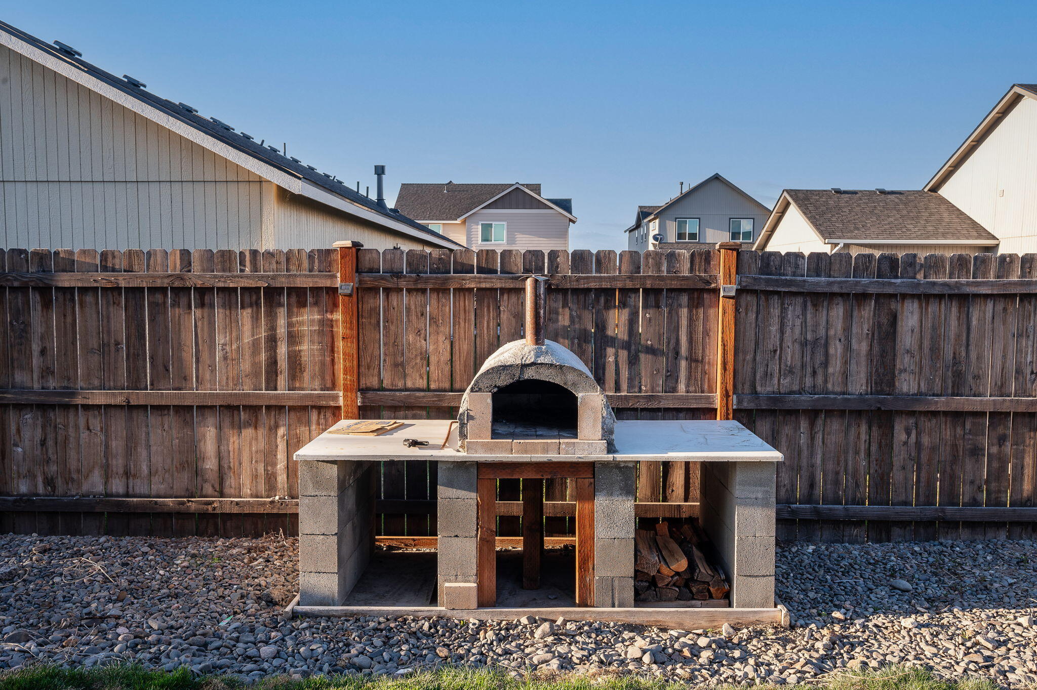 Backyard masonry pizza oven with wood storage, gravel ground cover, and a tall wooden privacy fence.