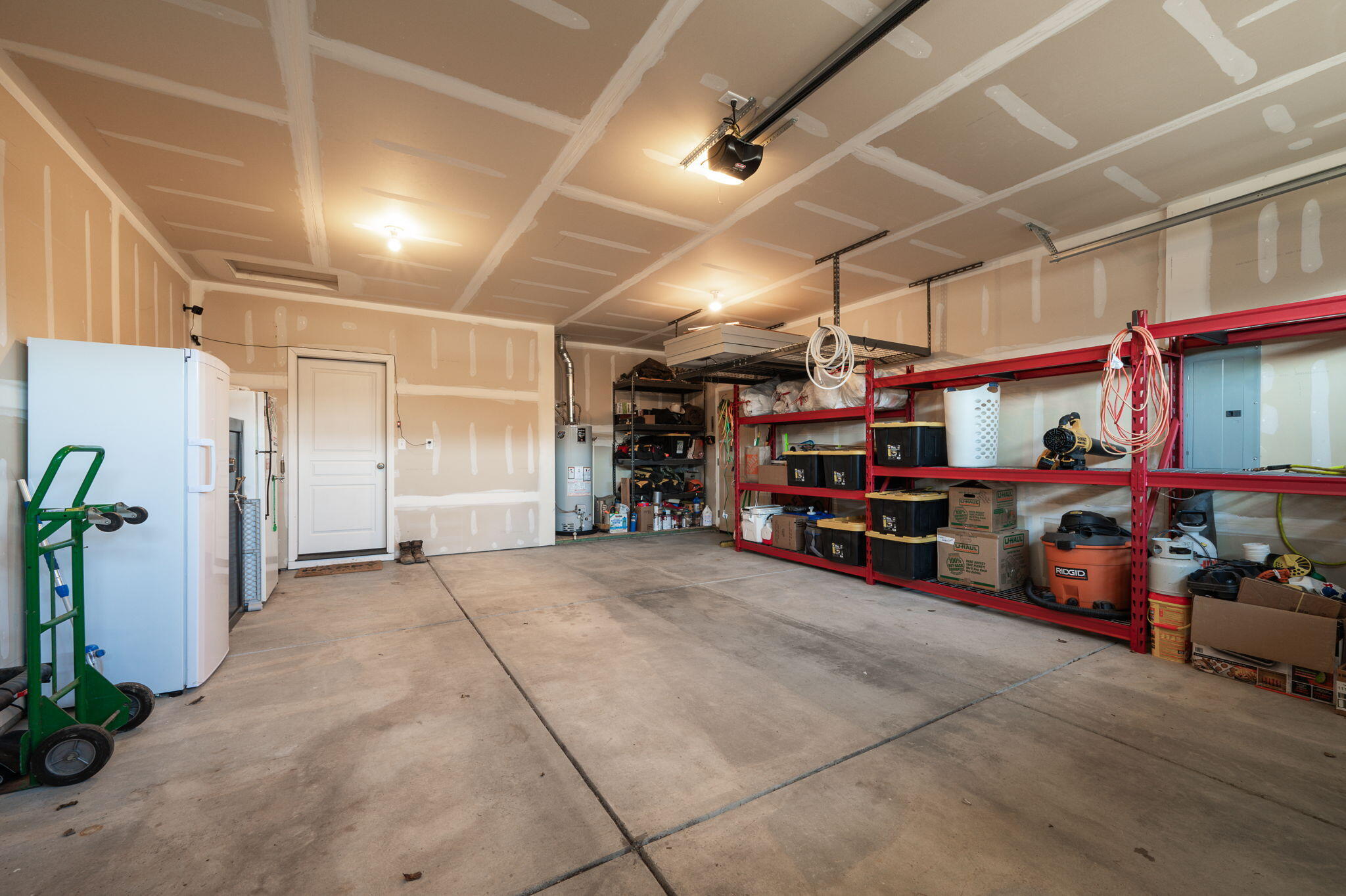 Spacious garage with red storage shelving, a concrete floor, refrigerator, and ceiling-mounted door opener.