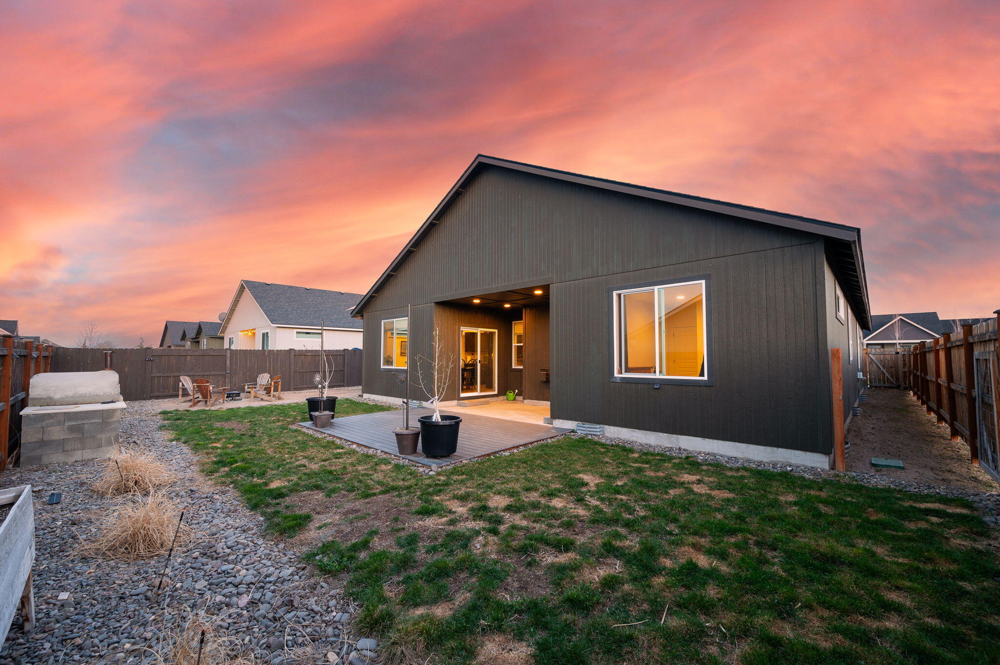Dark-sided home with a covered patio and fenced backyard beneath a pink sunset sky.