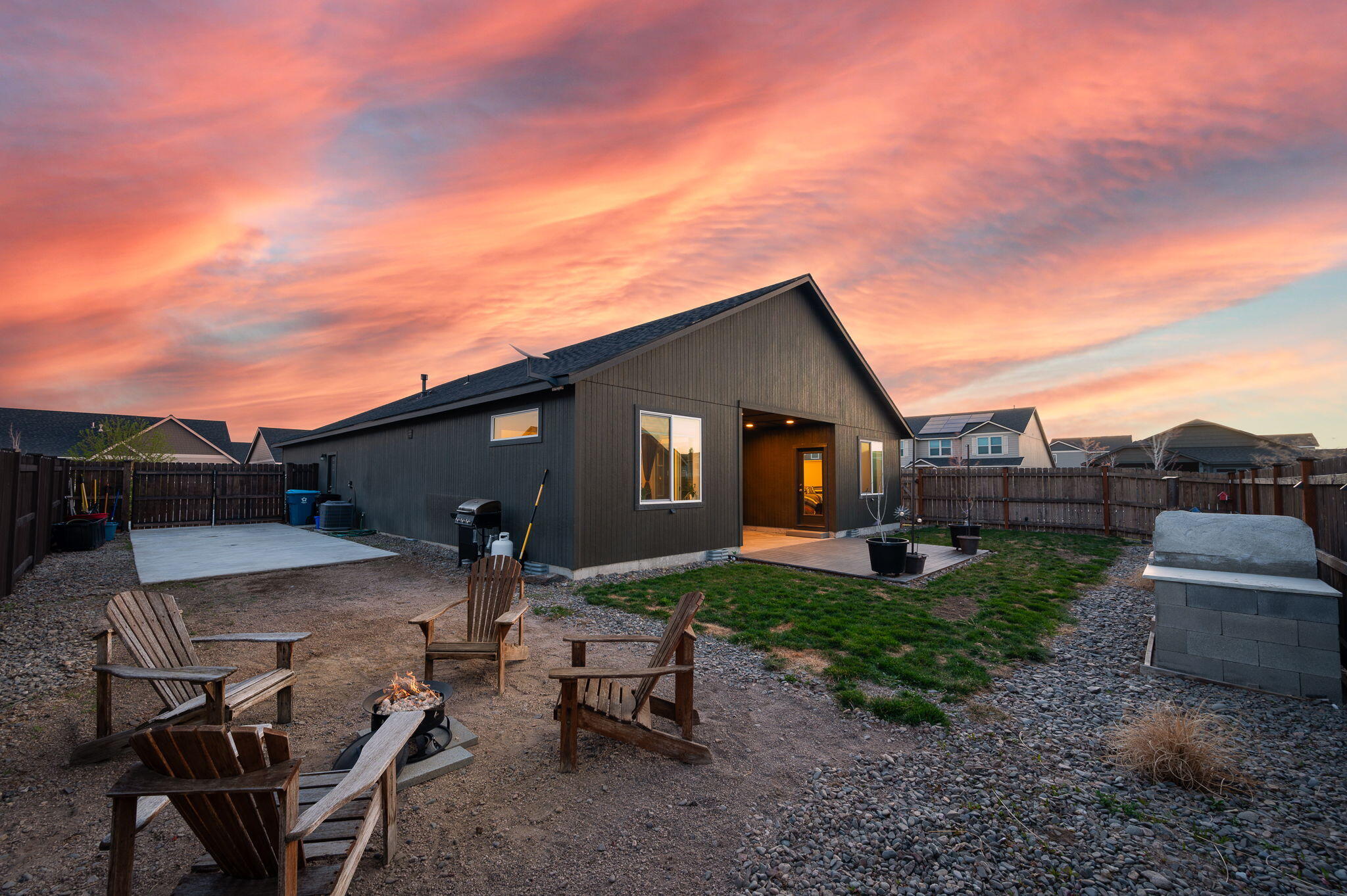 Dark-sided home and fenced backyard with patio seating and a fire pit at dusk.