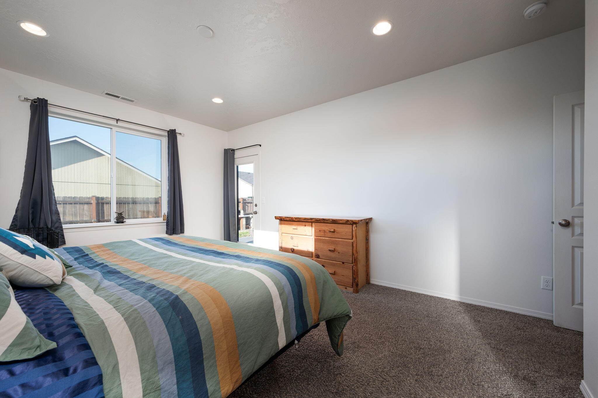 Bedroom with a striped comforter, wooden dresser, and large curtained window.