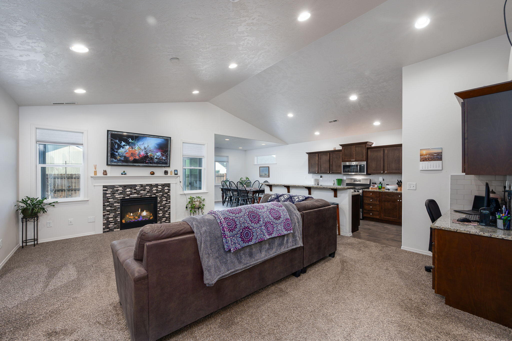 Open-plan living room with vaulted ceiling, tiled fireplace, brown sofas, and kitchen beyond.