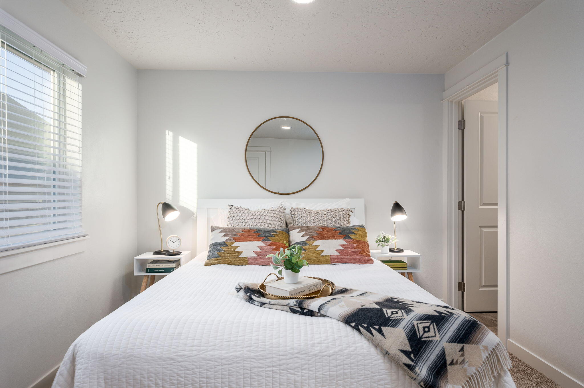 Staged bedroom with a white bed, patterned pillows, a round mirror, and daylight through window blinds.