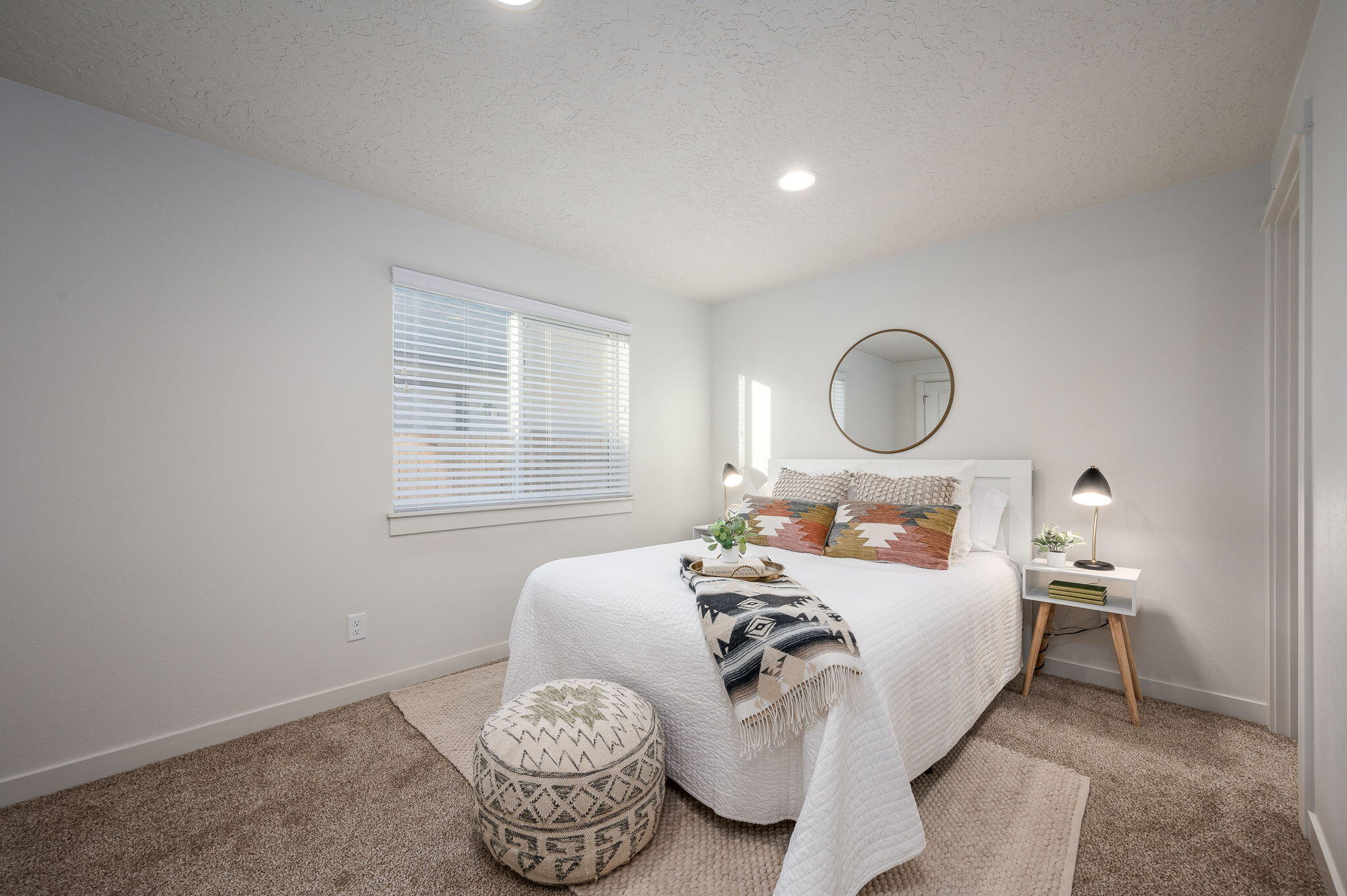 Staged bedroom with a white bed, patterned pillows and throw, and a round mirror.
