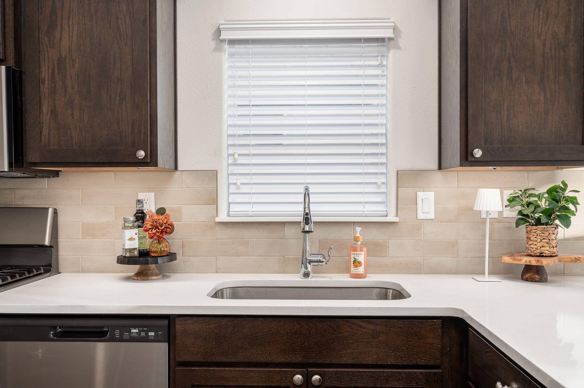 Kitchen sink beneath a window with white blinds, dark wood cabinets, and a white countertop.