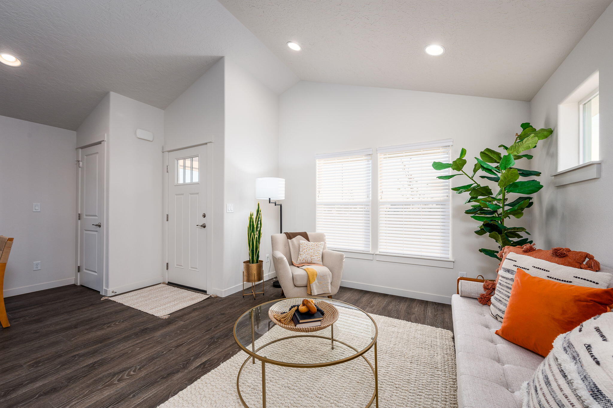 Staged living room with vaulted ceiling, orange sofa cushions, round glass coffee table, and large windows.