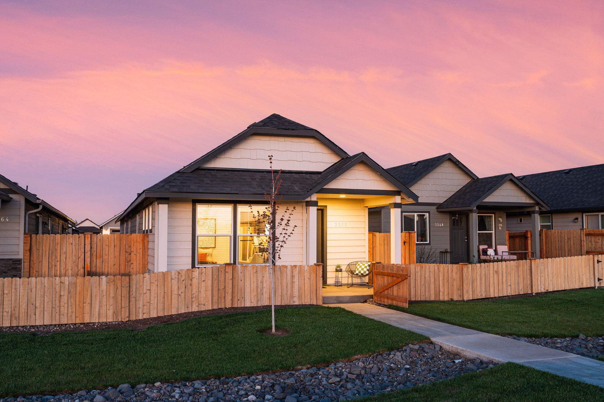 Single-story home with a fenced lawn and warmly lit covered entry beneath a pink dusk sky.