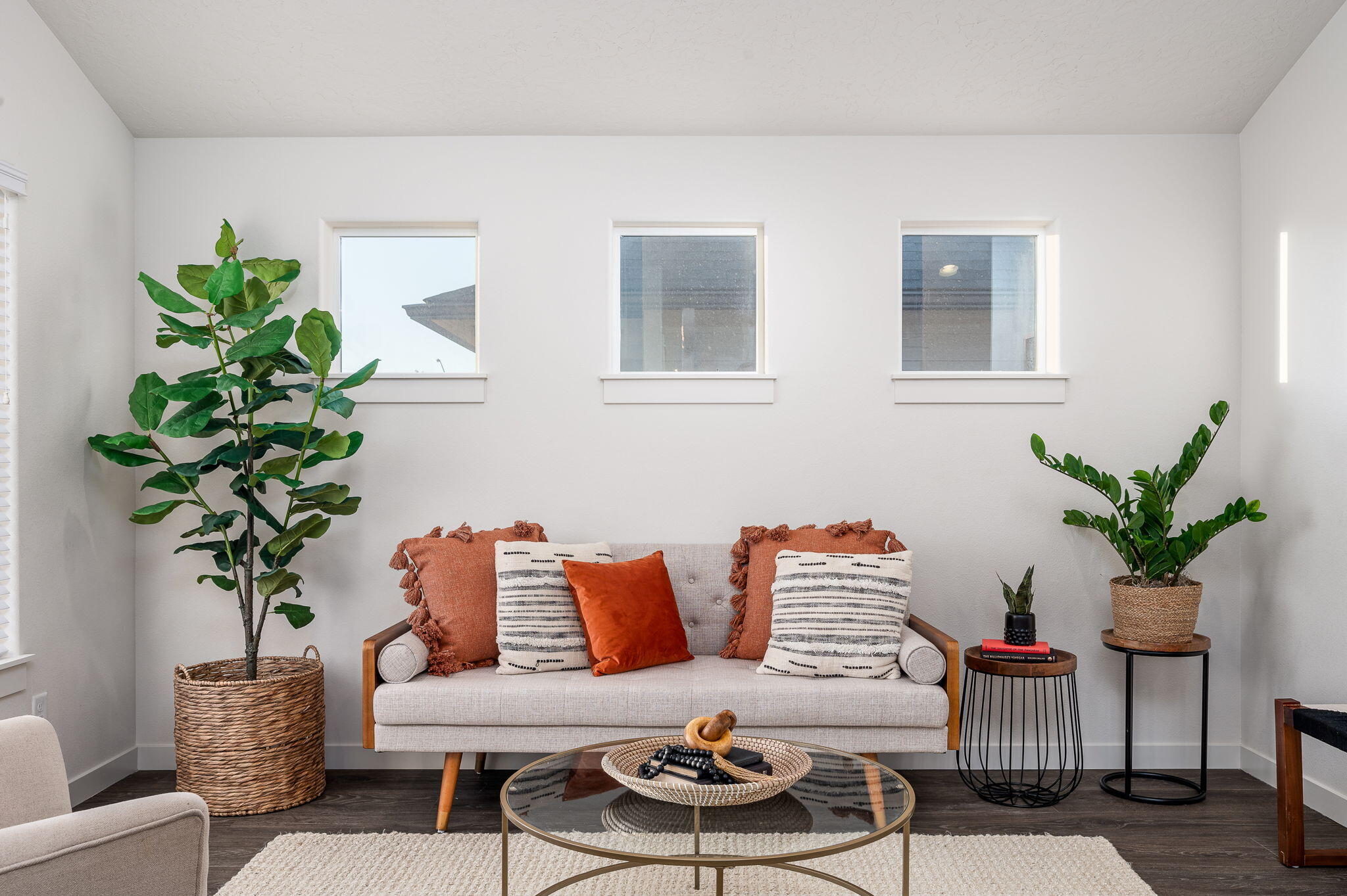 Staged living room with a gray sofa, rust-colored pillows, tall plants, and three high windows.