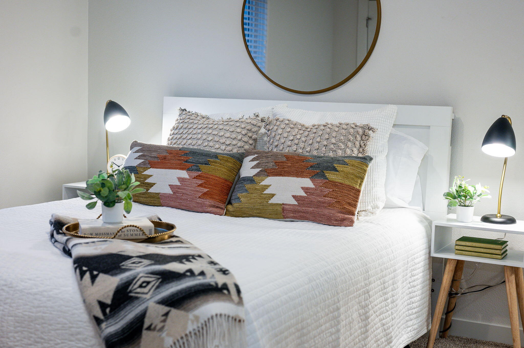 Staged bedroom with a white bed, colorful geometric pillows, patterned throw, and round wall mirror.