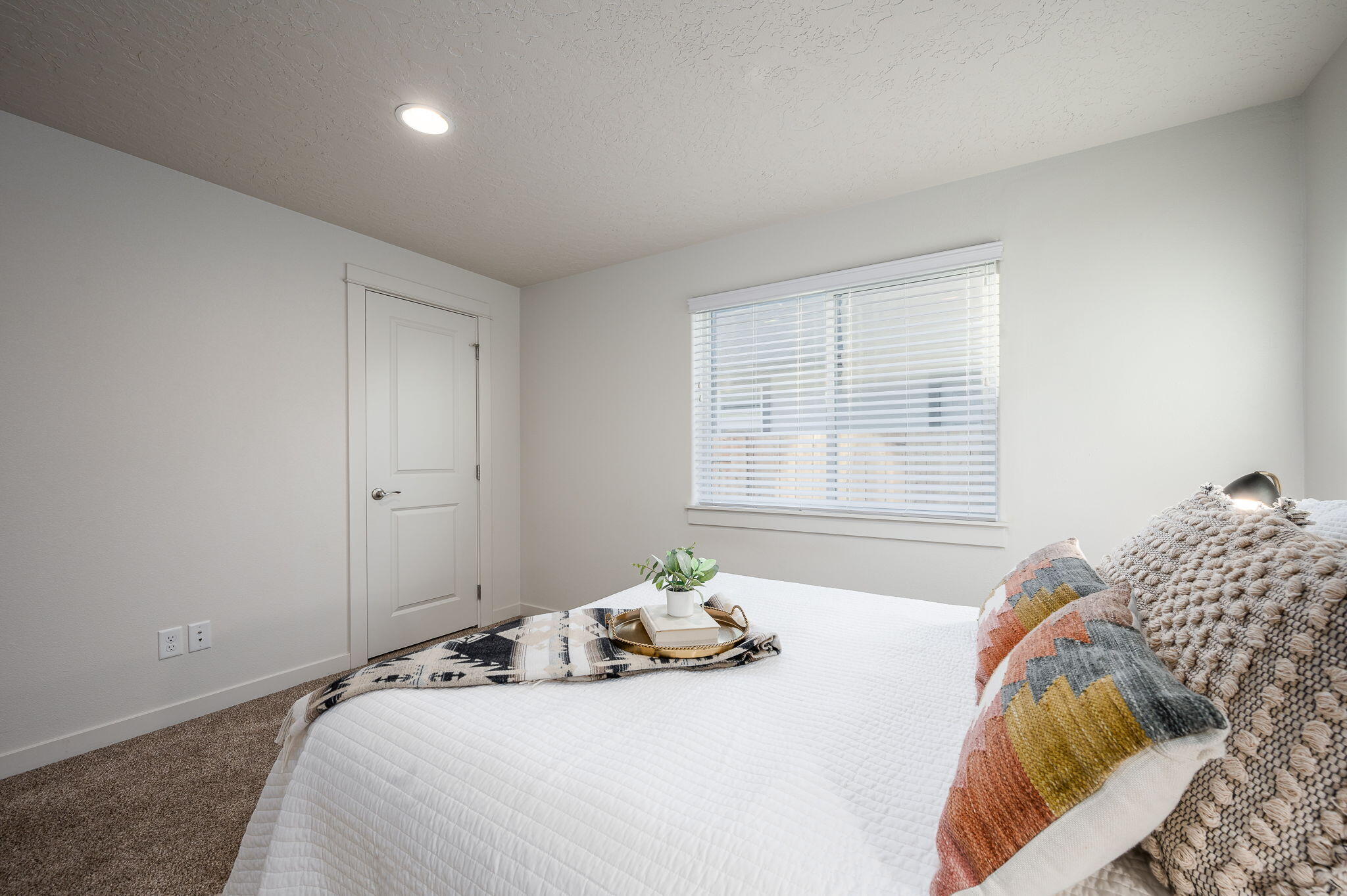 Staged bedroom with a white quilted bed, colorful pillows, and a window with white blinds.