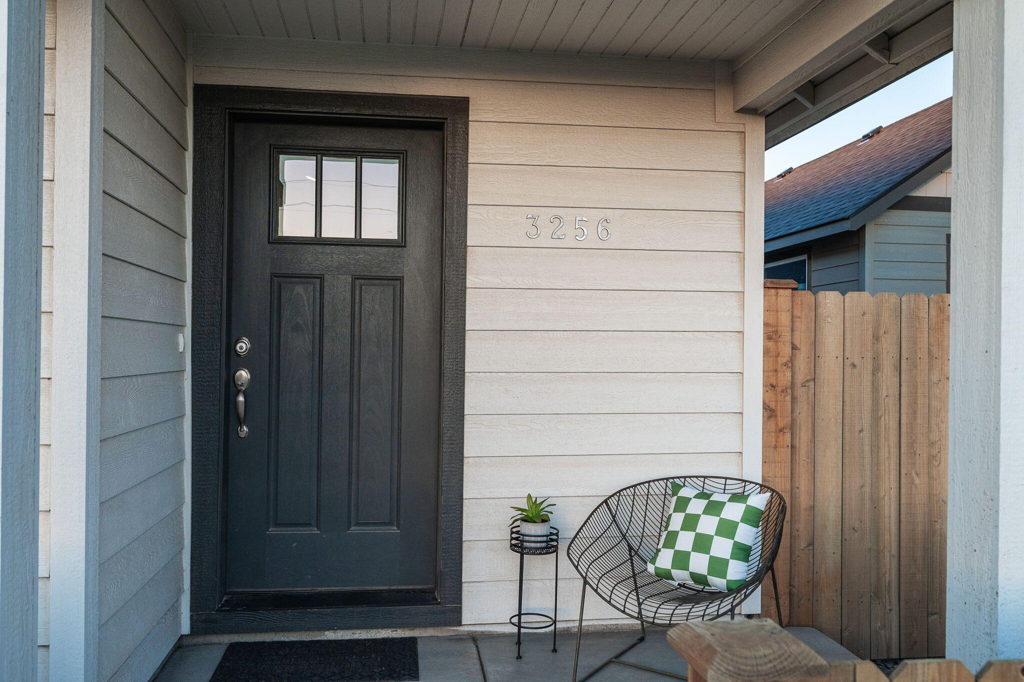Home’s covered entry porch with black front door, address 3256, and wire chair beside a wooden fence.