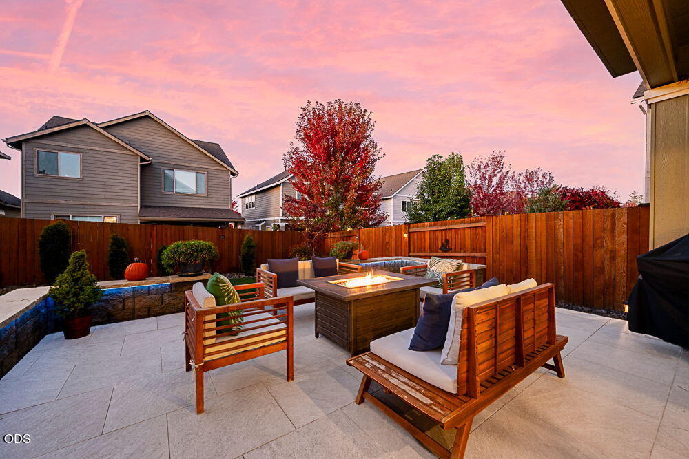Backyard patio with cushioned wood seating around a lit fire table and a tall privacy fence.