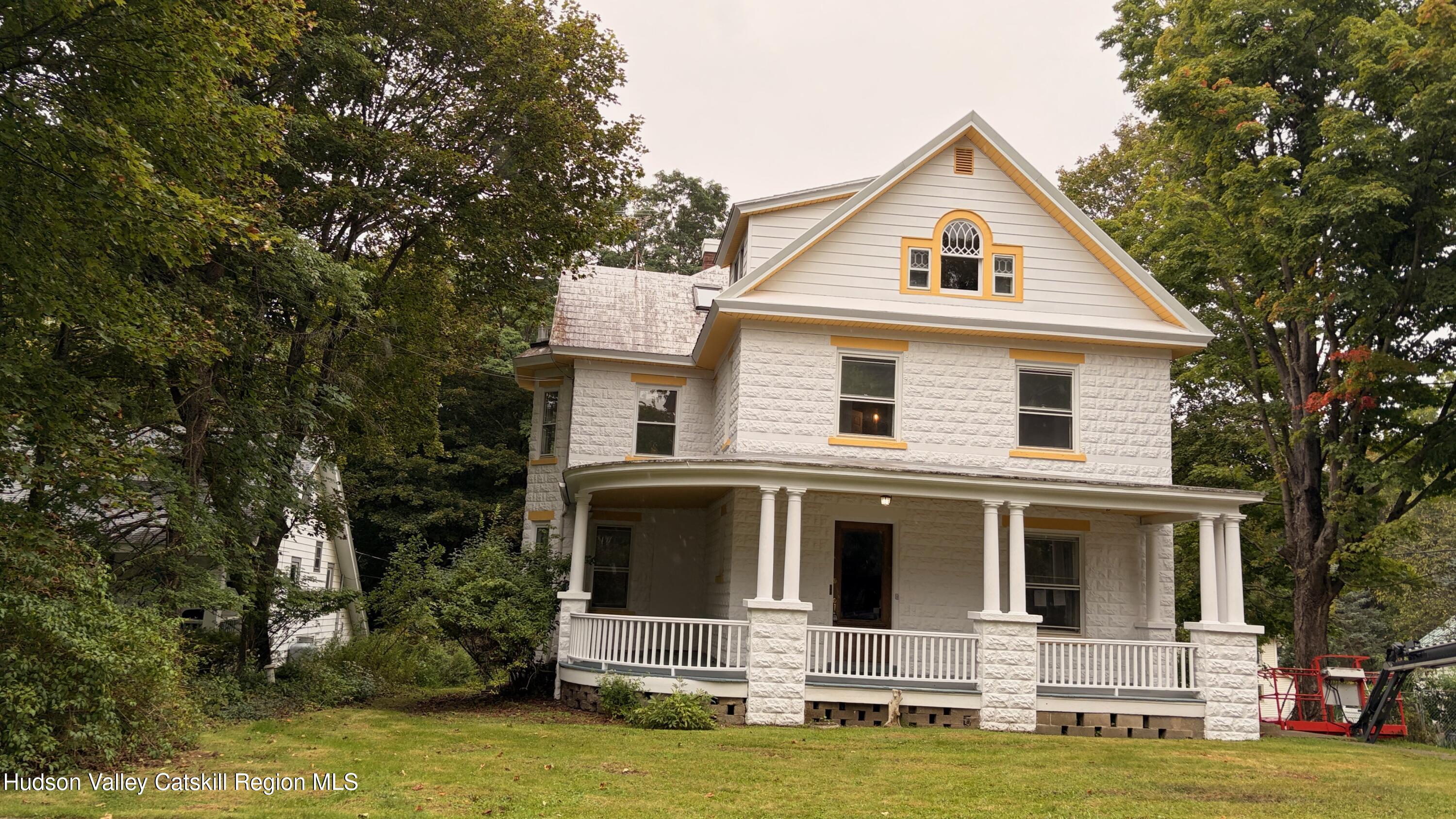 White stone house with a columned wraparound porch and decorative gable windows, surrounded by mature trees.