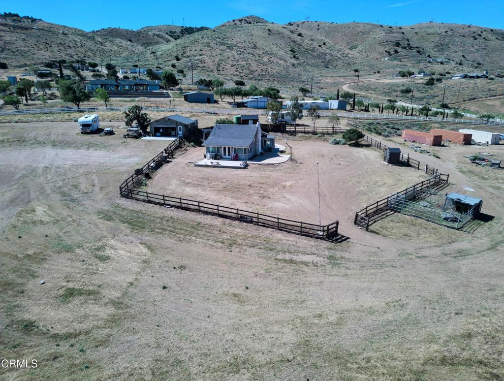 Aerial view of a rural house and fenced yard with outbuildings and rolling hills beyond.