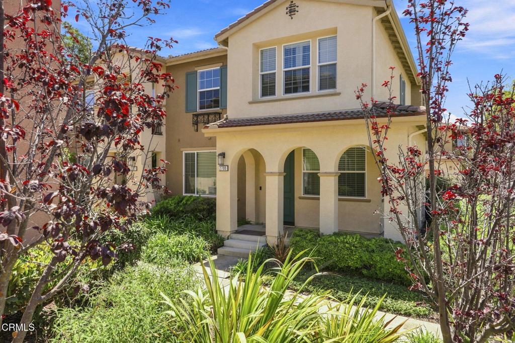 Two-story beige stucco home with an arched porch, green shutters, and leafy landscaped front yard.