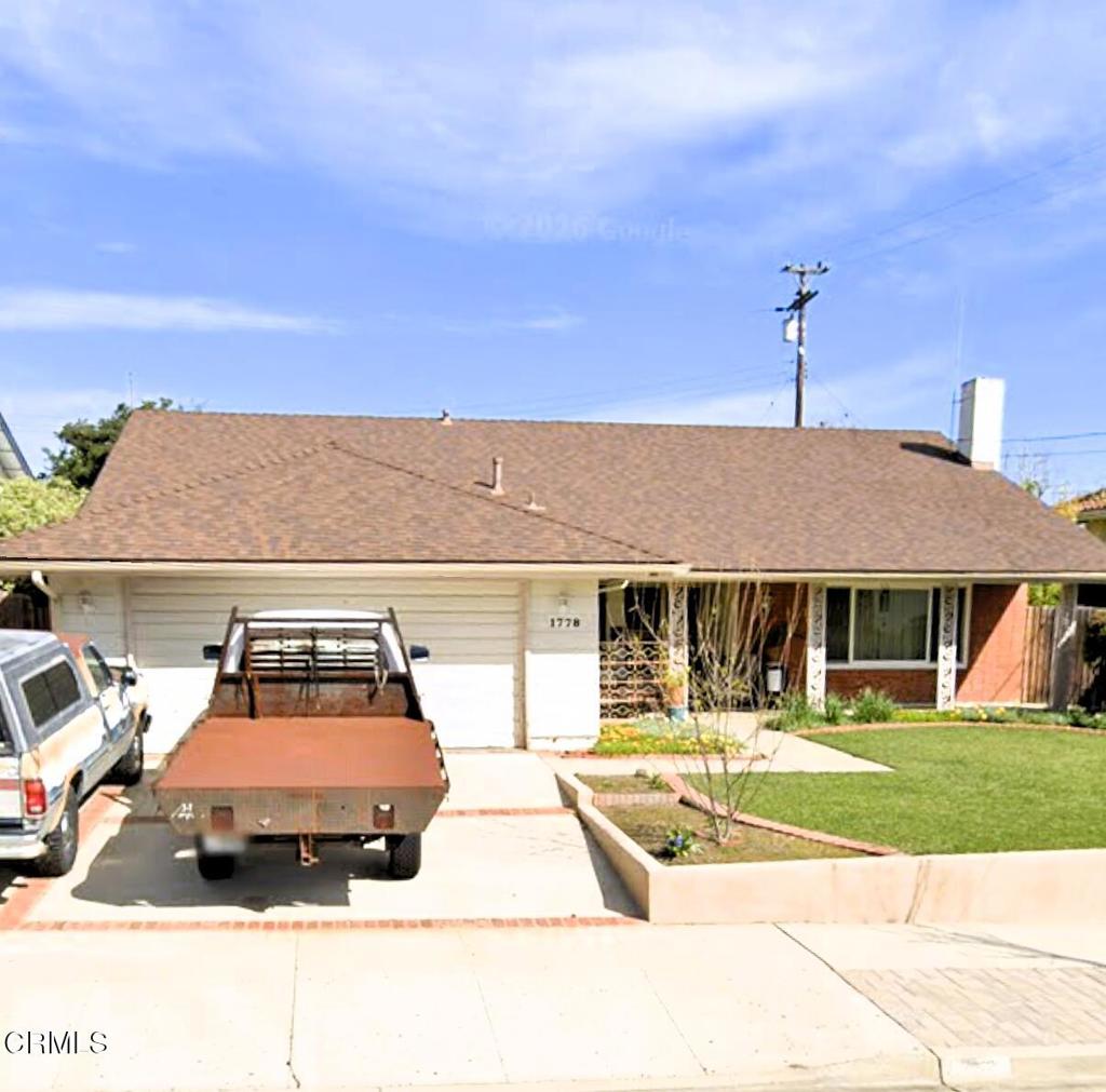 Single-story house with a broad shingled roof, attached garage, driveway vehicles, and front lawn.