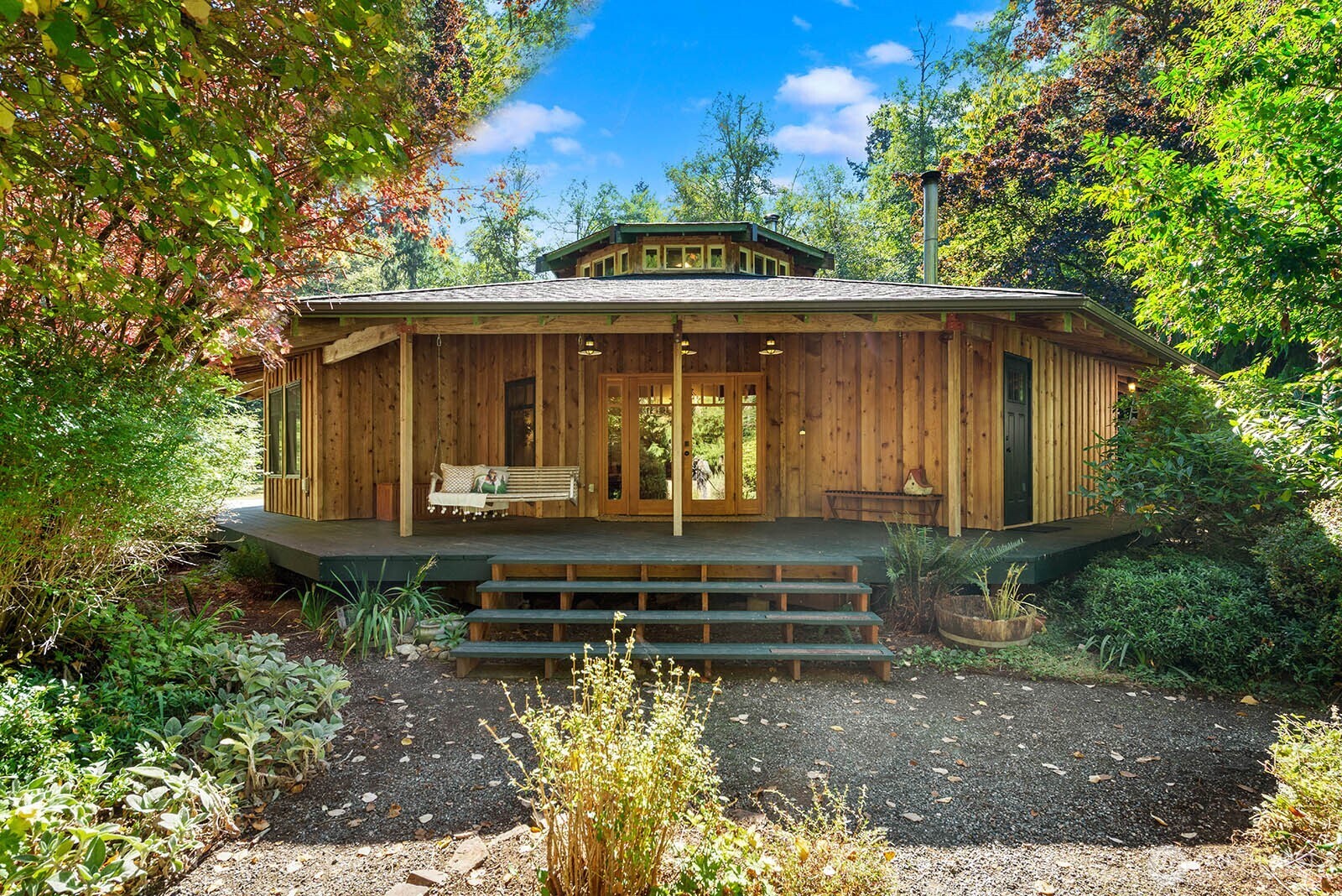 Wood-sided home with a broad covered porch and steps, surrounded by mature trees and garden landscaping.