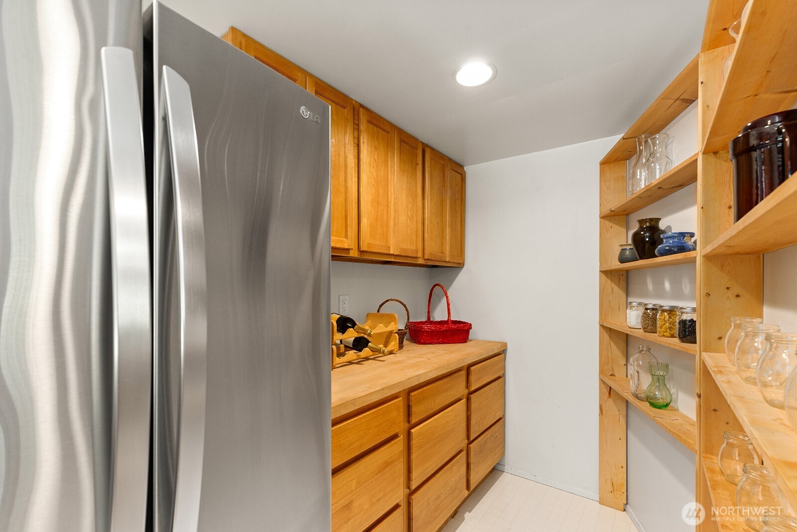 Pantry with warm wood cabinetry, a stainless steel refrigerator, and open shelves of jars and glassware.