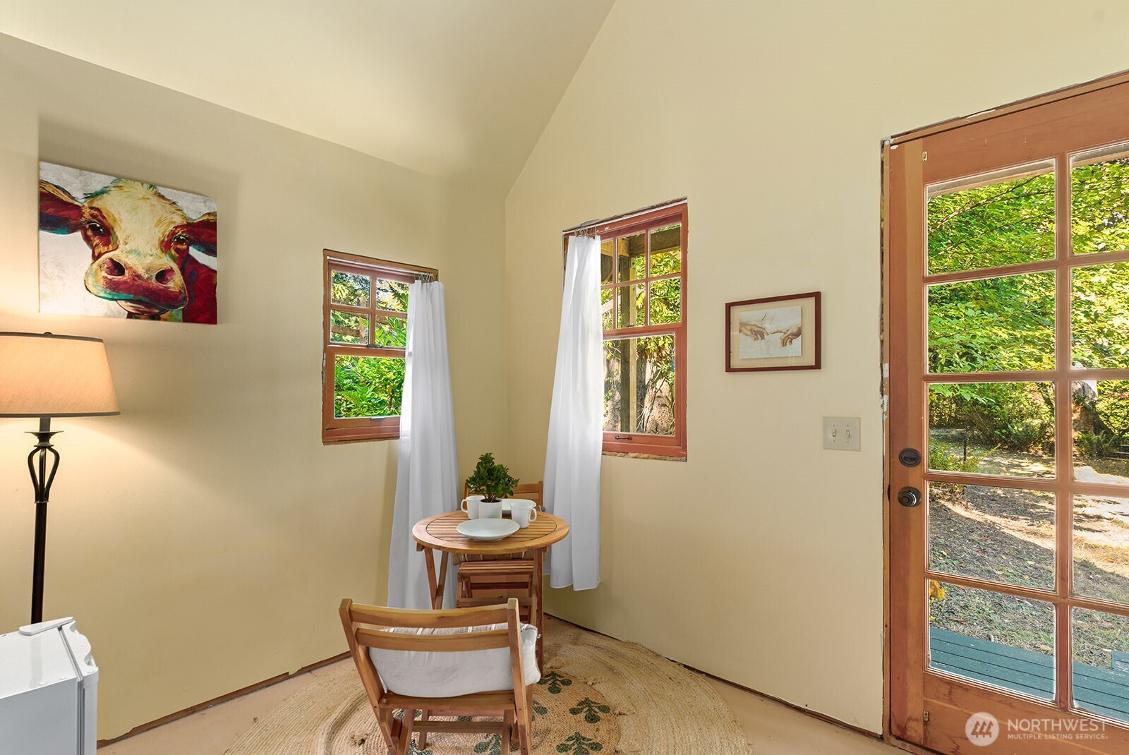 Breakfast nook with a round wooden table, cow artwork, and a glass door overlooking a leafy yard.