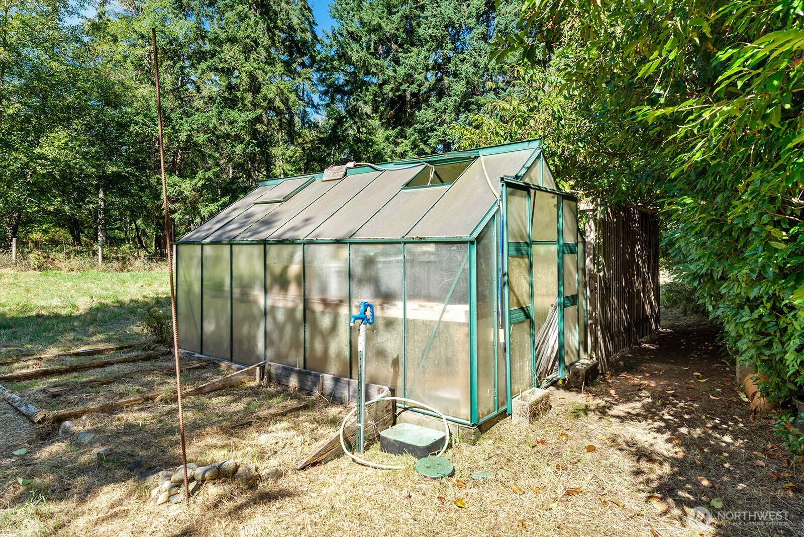 Green-framed glass greenhouse among trees, with raised garden beds and a blue water pump.