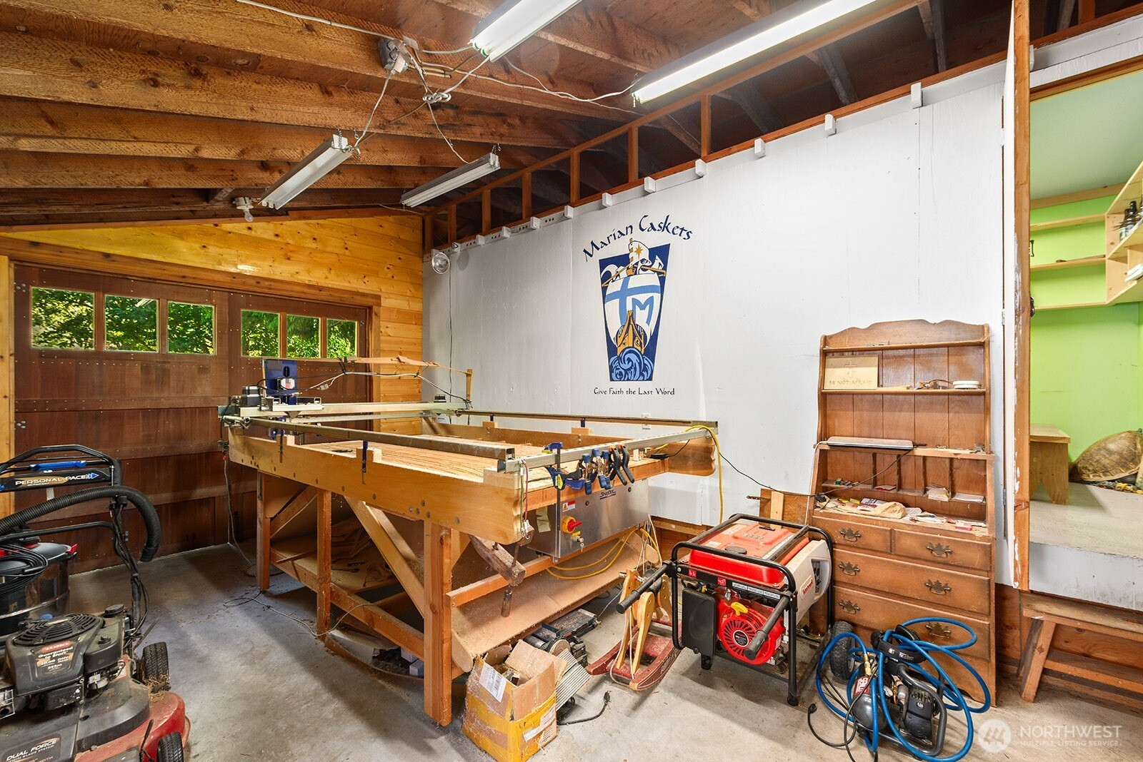 Workshop with a large wooden workbench, woodworking machinery, and a Marian Caskets sign.