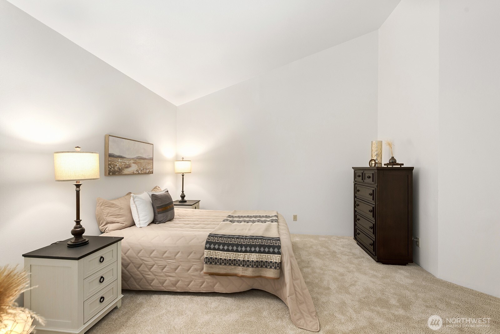 Staged bedroom with a quilted bed, patterned throw, tall dresser, and beige carpet.