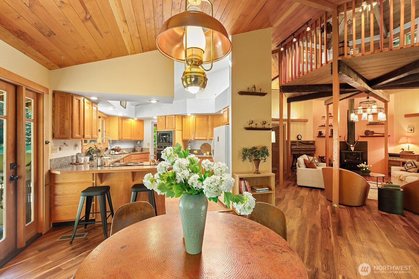 Open-plan kitchen and living area with wood cabinetry, a timber ceiling, and an upstairs loft railing.