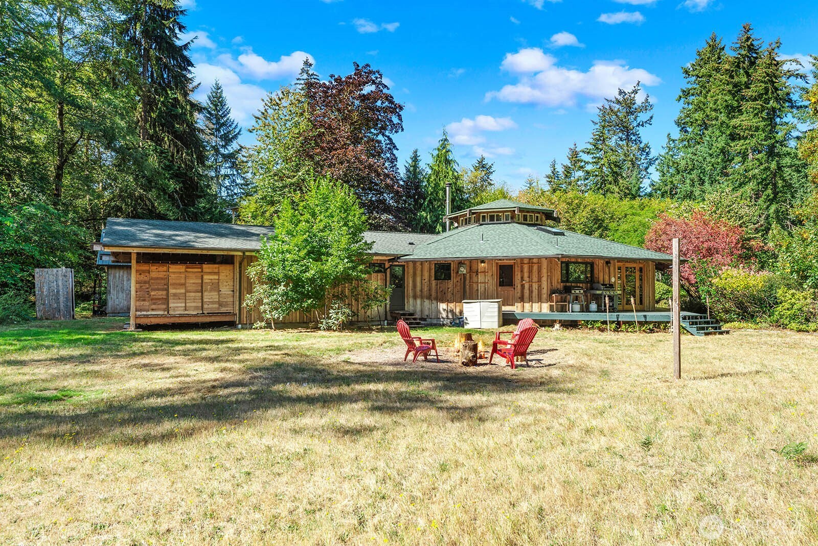 Wood-sided home with a raised cupola, surrounded by trees; red chairs sit around a fire pit.
