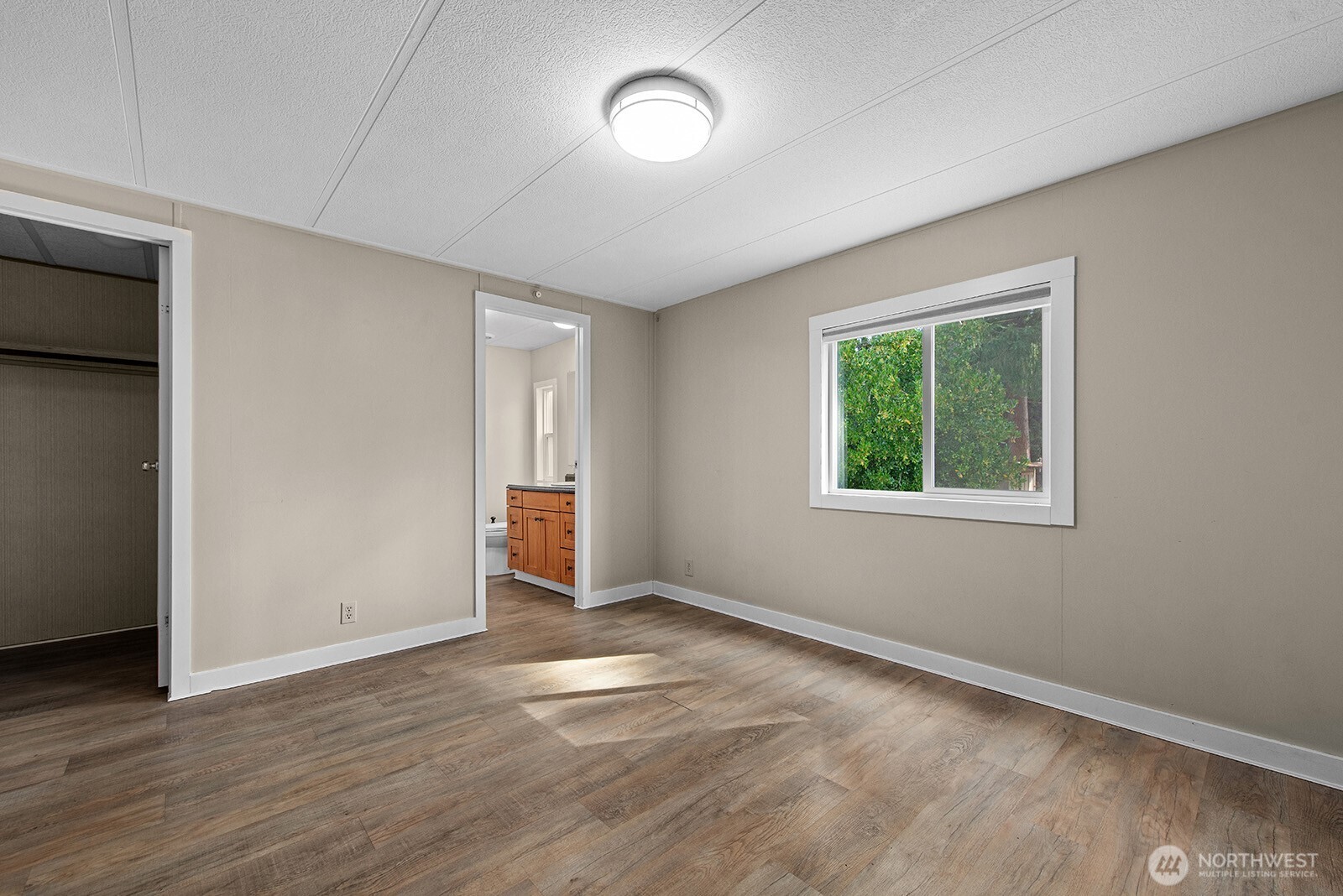 Empty bedroom with wood-look flooring, a window overlooking greenery, and a closet doorway.