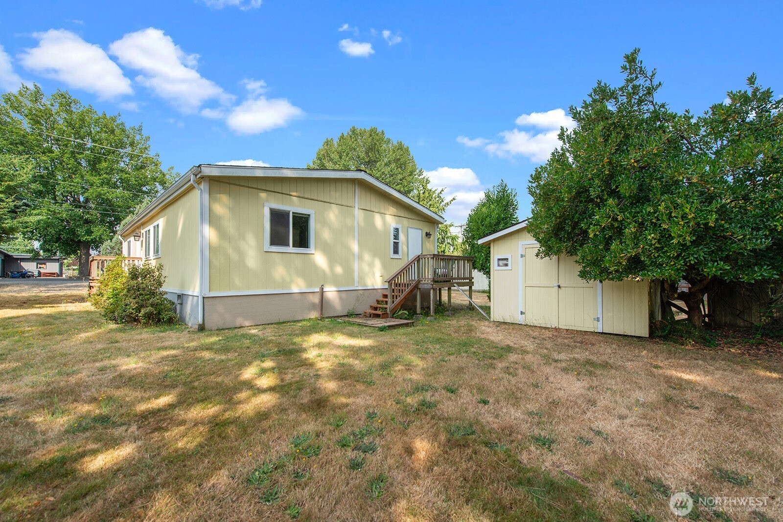 Pale yellow single-story home with a raised deck, detached shed, and dry grassy yard.