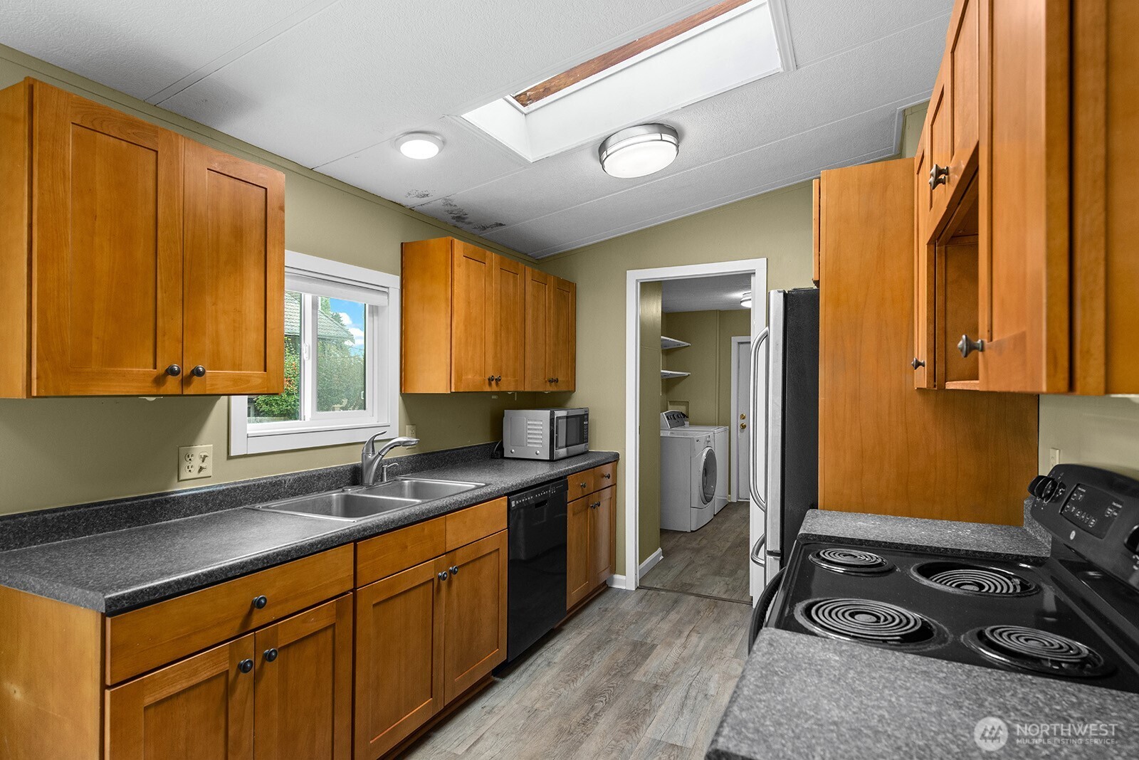 Kitchen with honey-colored cabinets, gray counters, skylight, and laundry area through doorway.