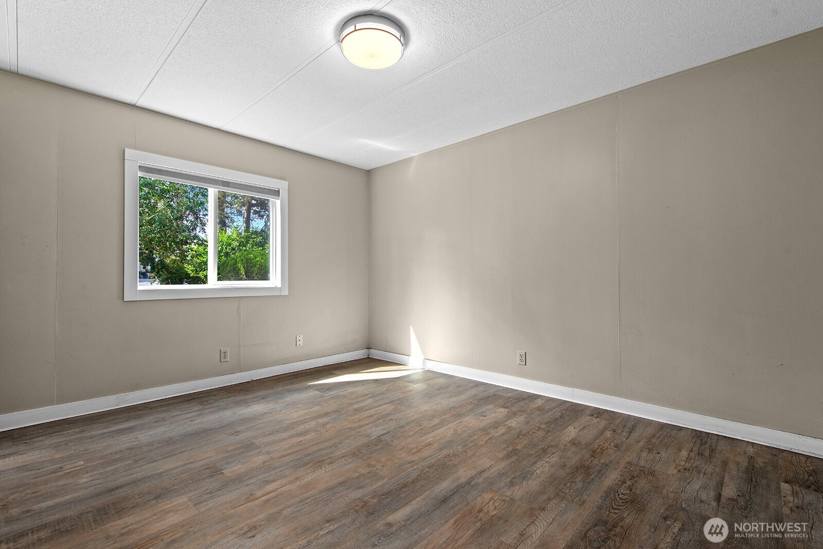 Empty room with wood-look flooring, beige walls, and a window overlooking greenery.
