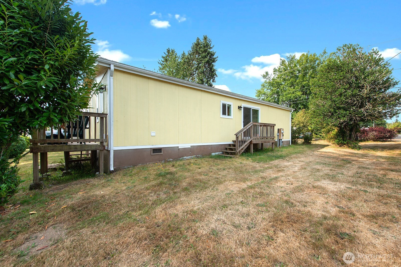 Pale yellow home with two wooden decks beside a dry lawn and leafy trees.