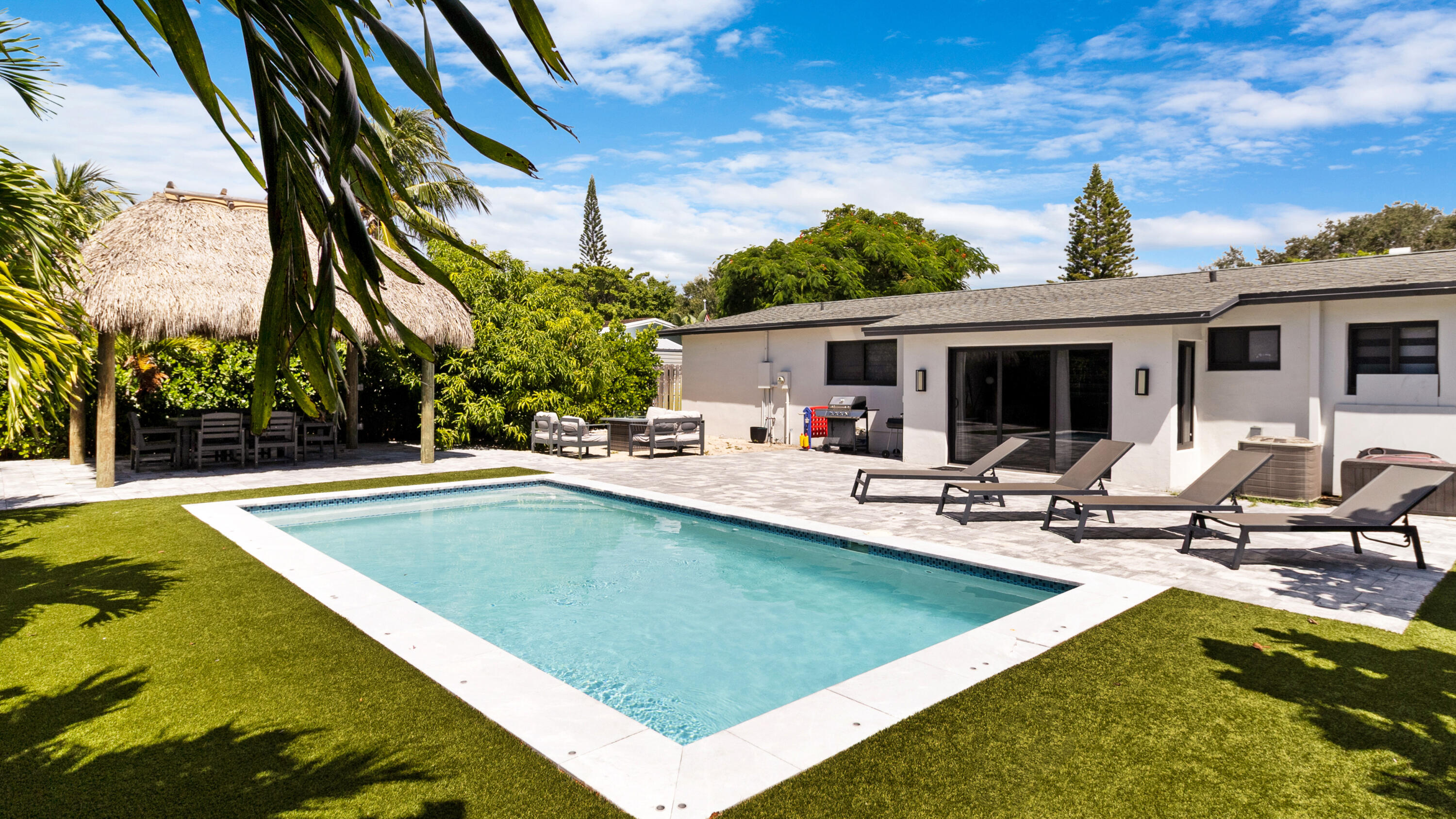 Backyard pool beside a modern home, with lounge chairs and a thatched-roof seating area.