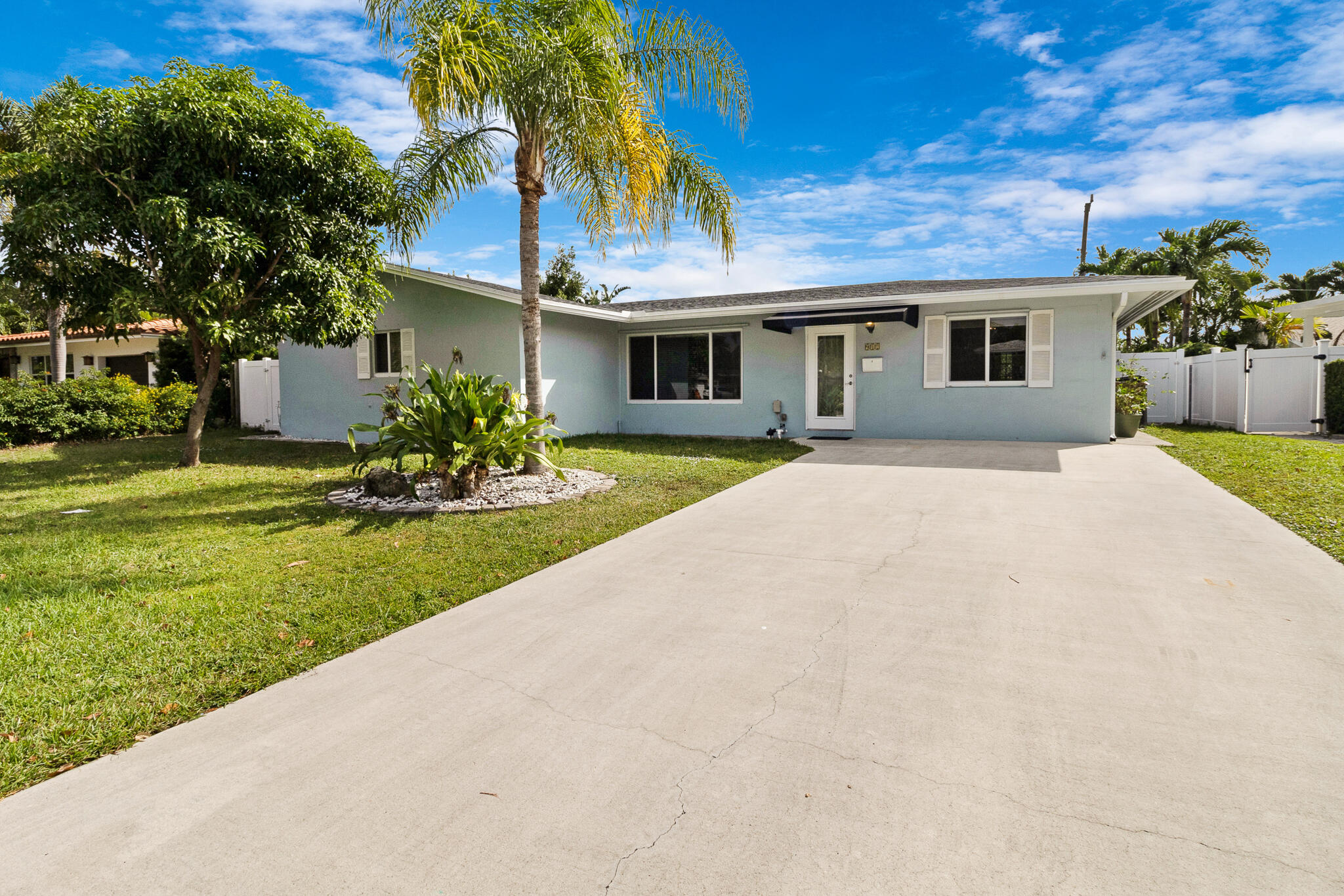 Single-story pale blue house with a wide driveway, fenced lawn, and palm trees.
