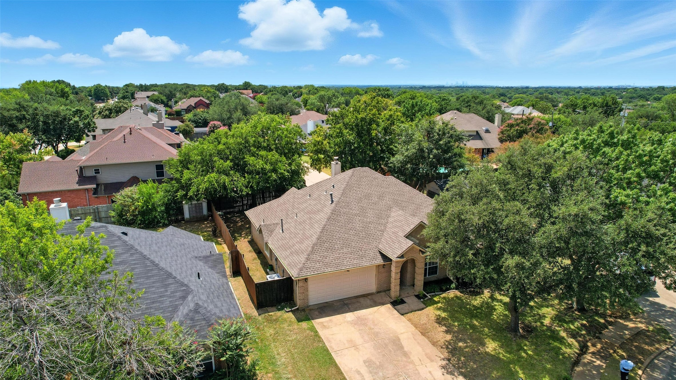 Aerial view of a brick home with a tan roof, attached garage, and tree-shaded driveway.