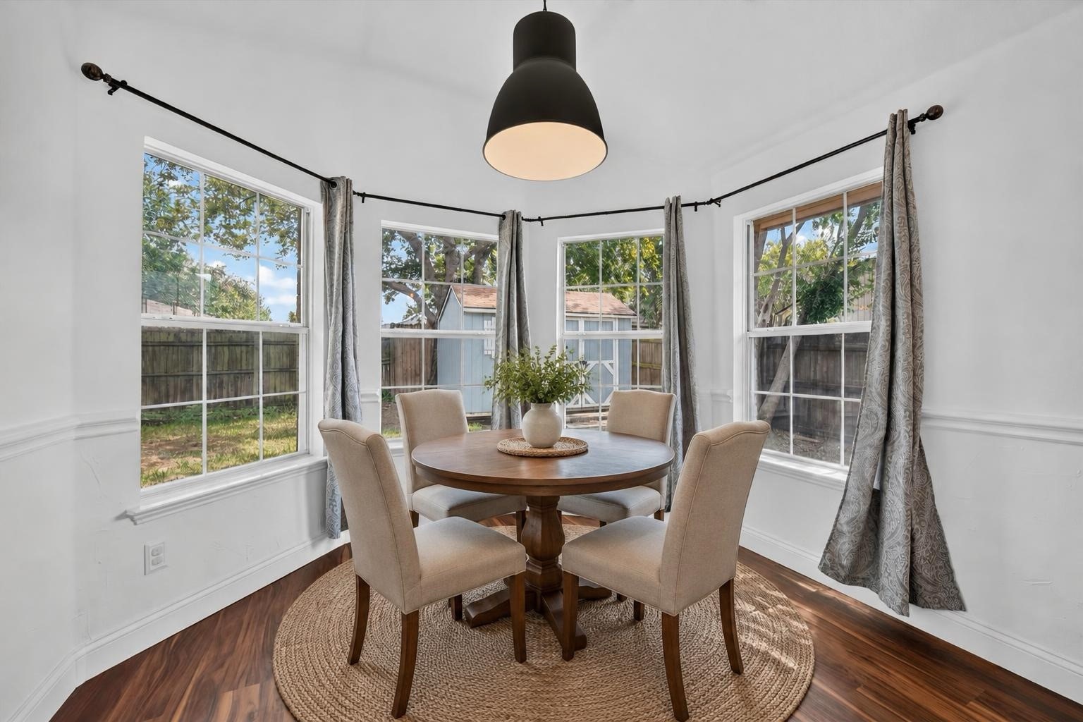 Dining room with a round wooden table, four upholstered chairs, and three large curtained windows.