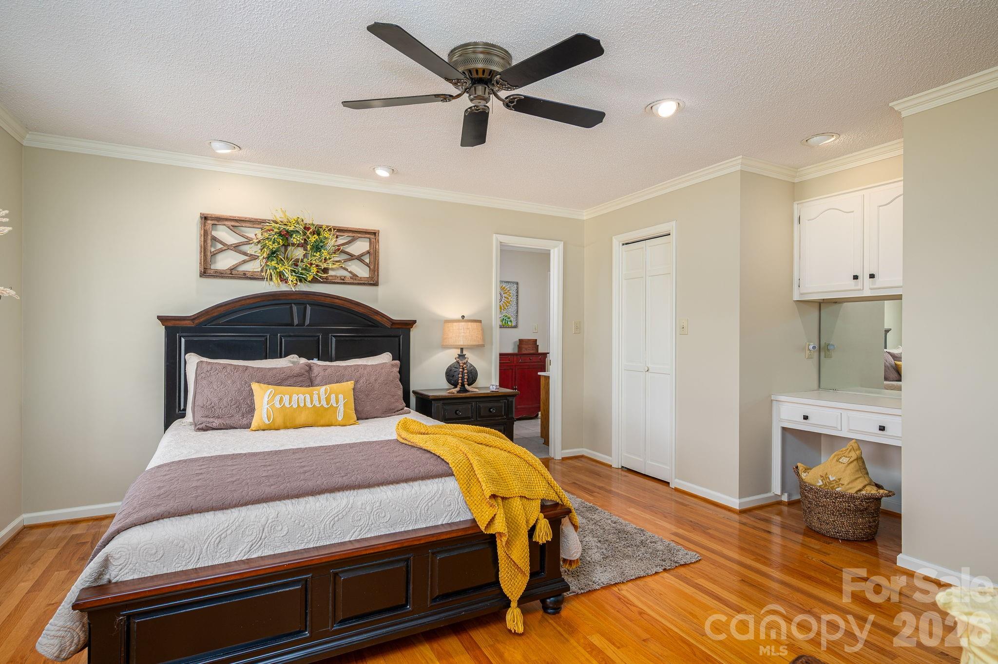 Staged bedroom with a dark wood bed, mustard yellow accents, hardwood floors, and a ceiling fan.