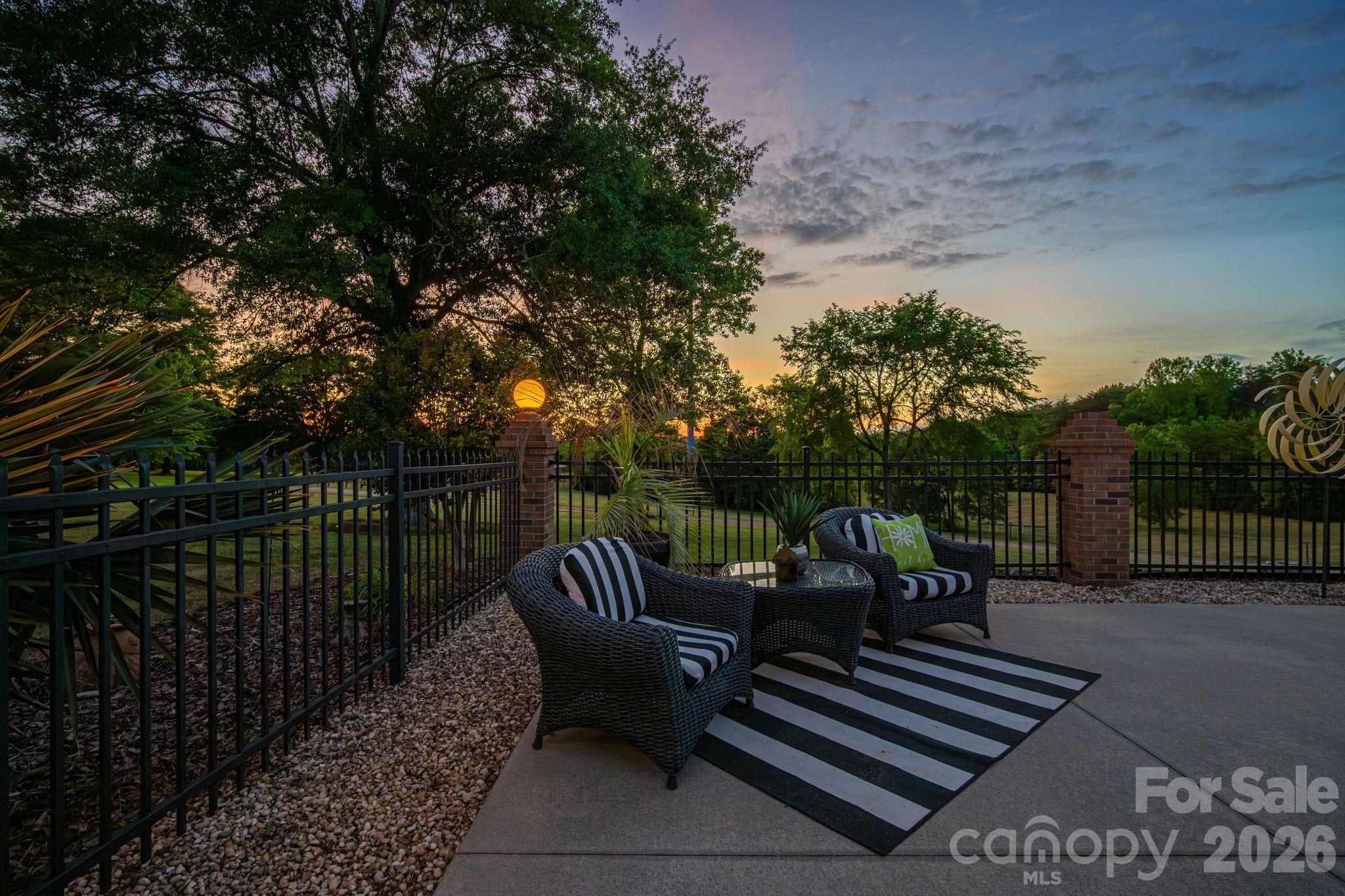 Patio seating with wicker chairs and a striped rug beside a fenced yard at sunset.
