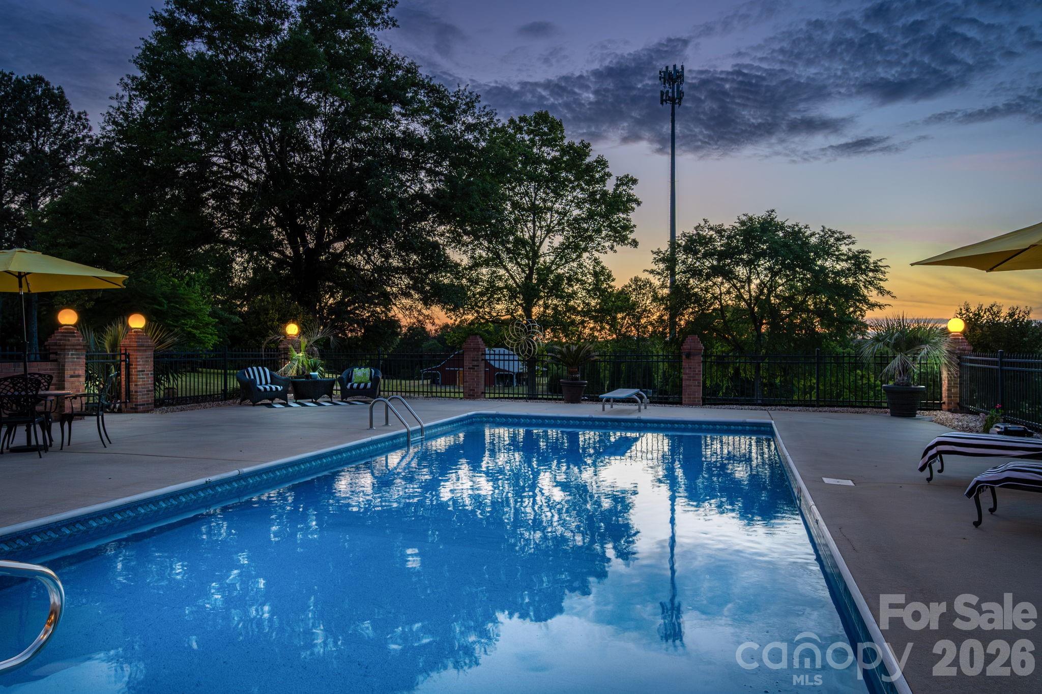 Backyard swimming pool and furnished patio at dusk, with illuminated brick posts and trees.