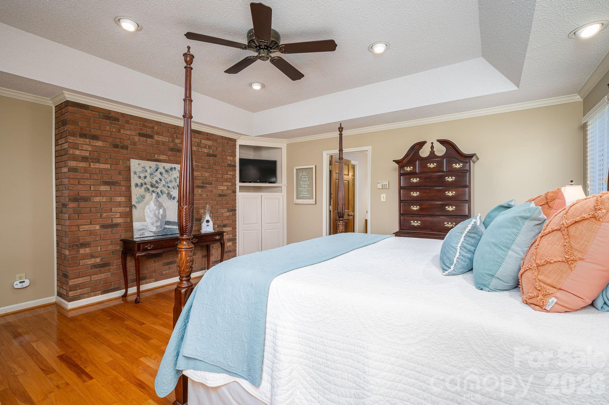 Furnished bedroom with a brick accent wall, hardwood floors, and a white bed with blue and coral pillows.