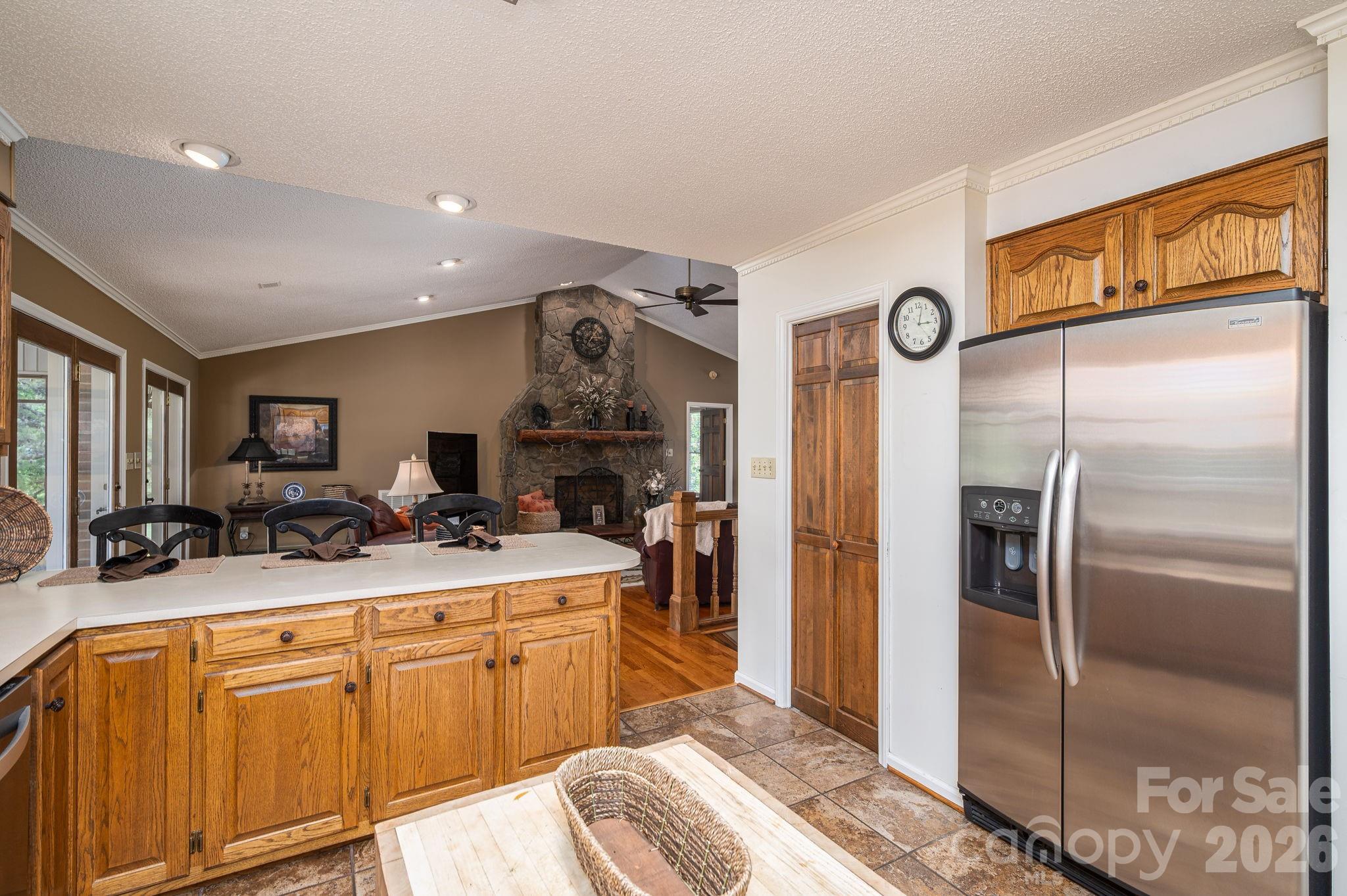 Kitchen with oak cabinetry and stainless refrigerator, opening to a living area with a stone fireplace.