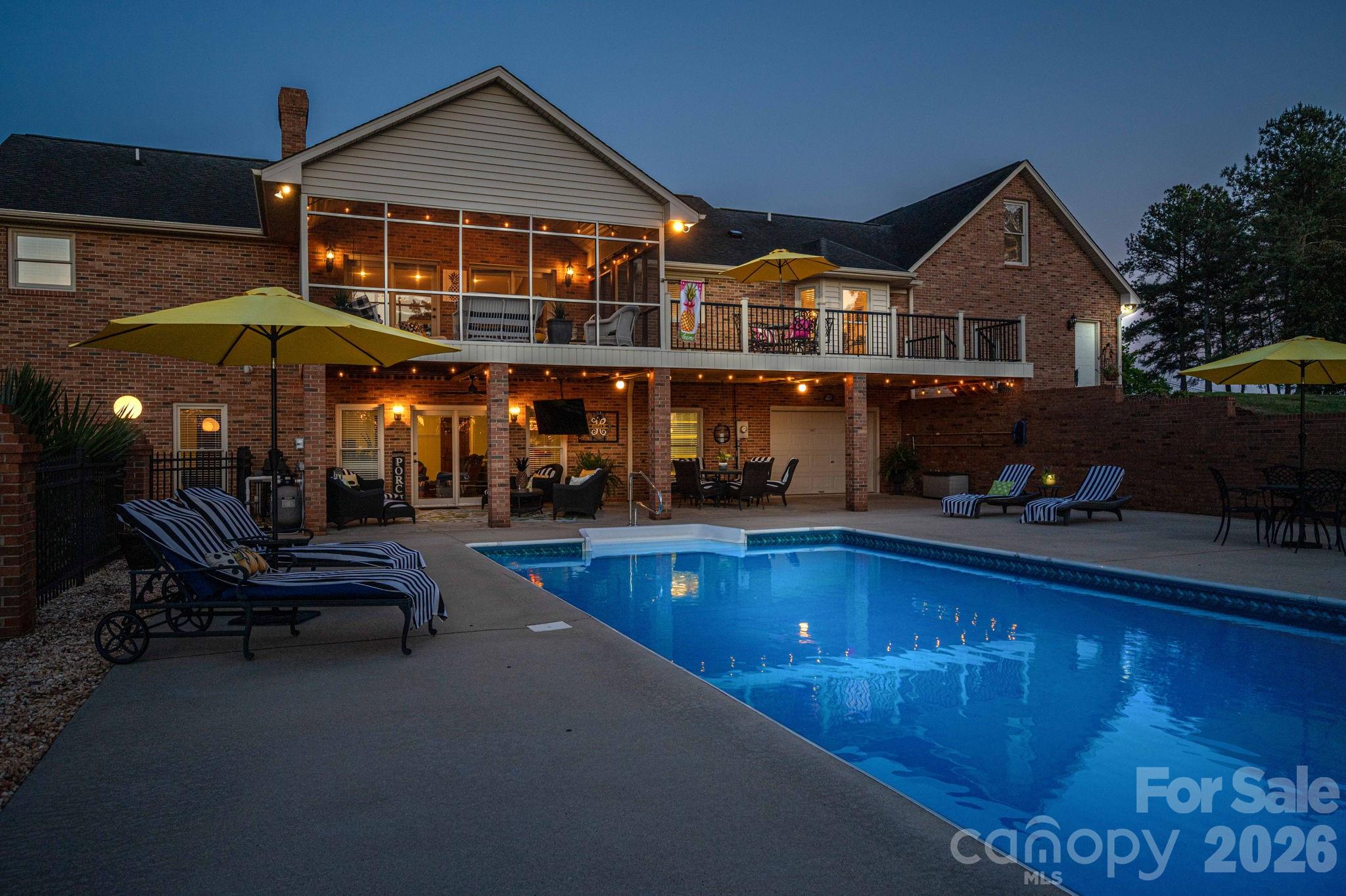 Brick house and two-level porch overlooking an illuminated swimming pool and furnished patio at dusk.