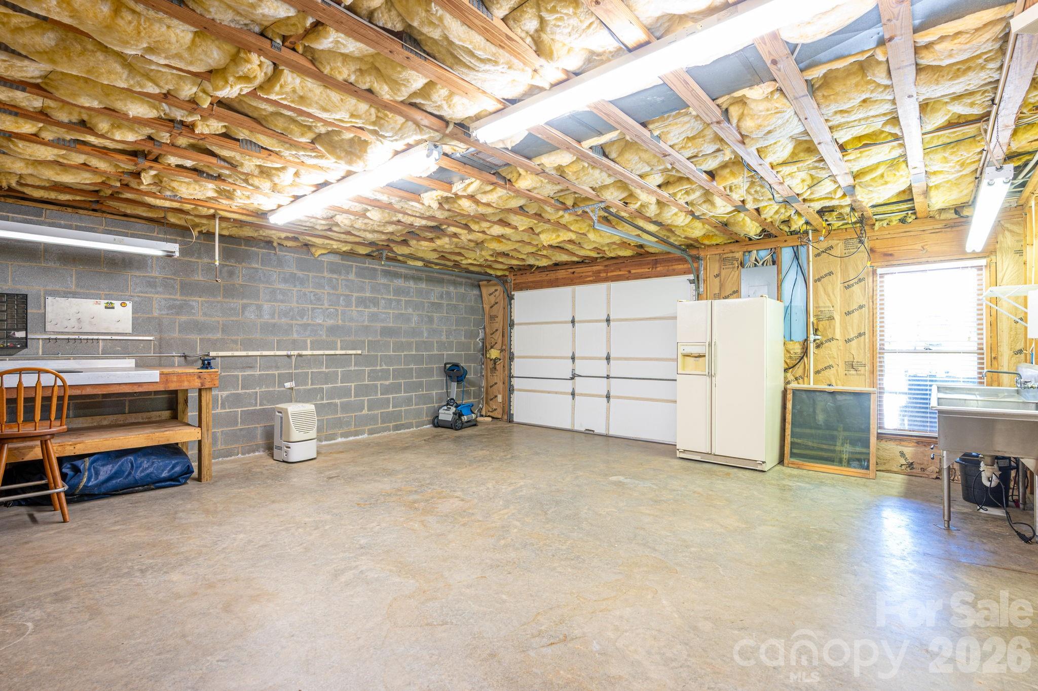 Spacious garage with exposed insulation, a workbench, refrigerator, and wide garage door.