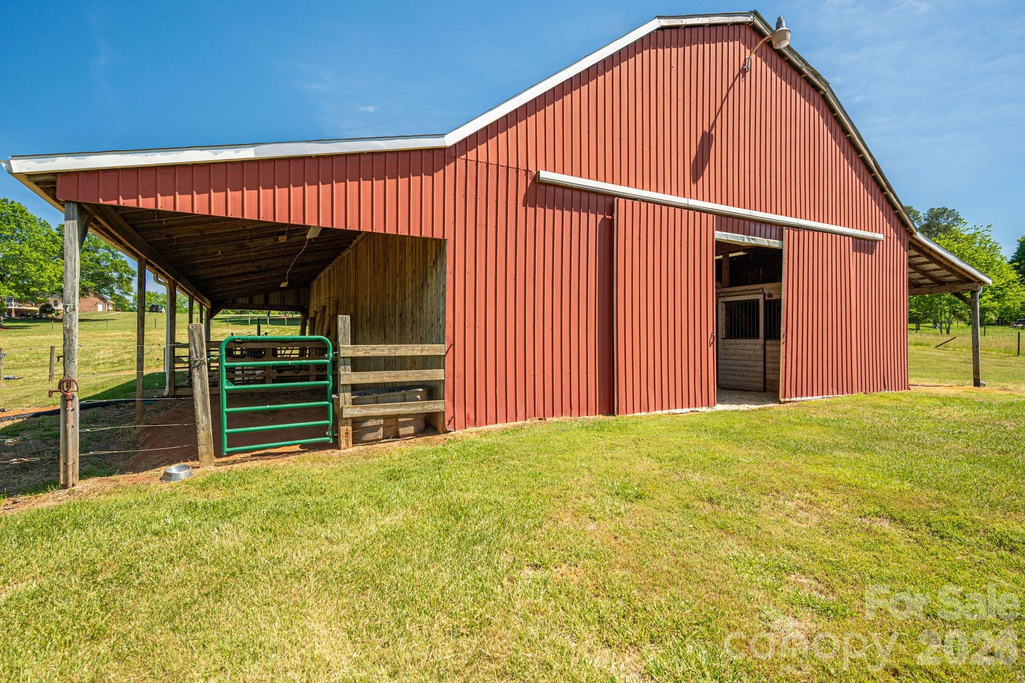 Red barn with a covered livestock area, wooden fencing, and a green gate.