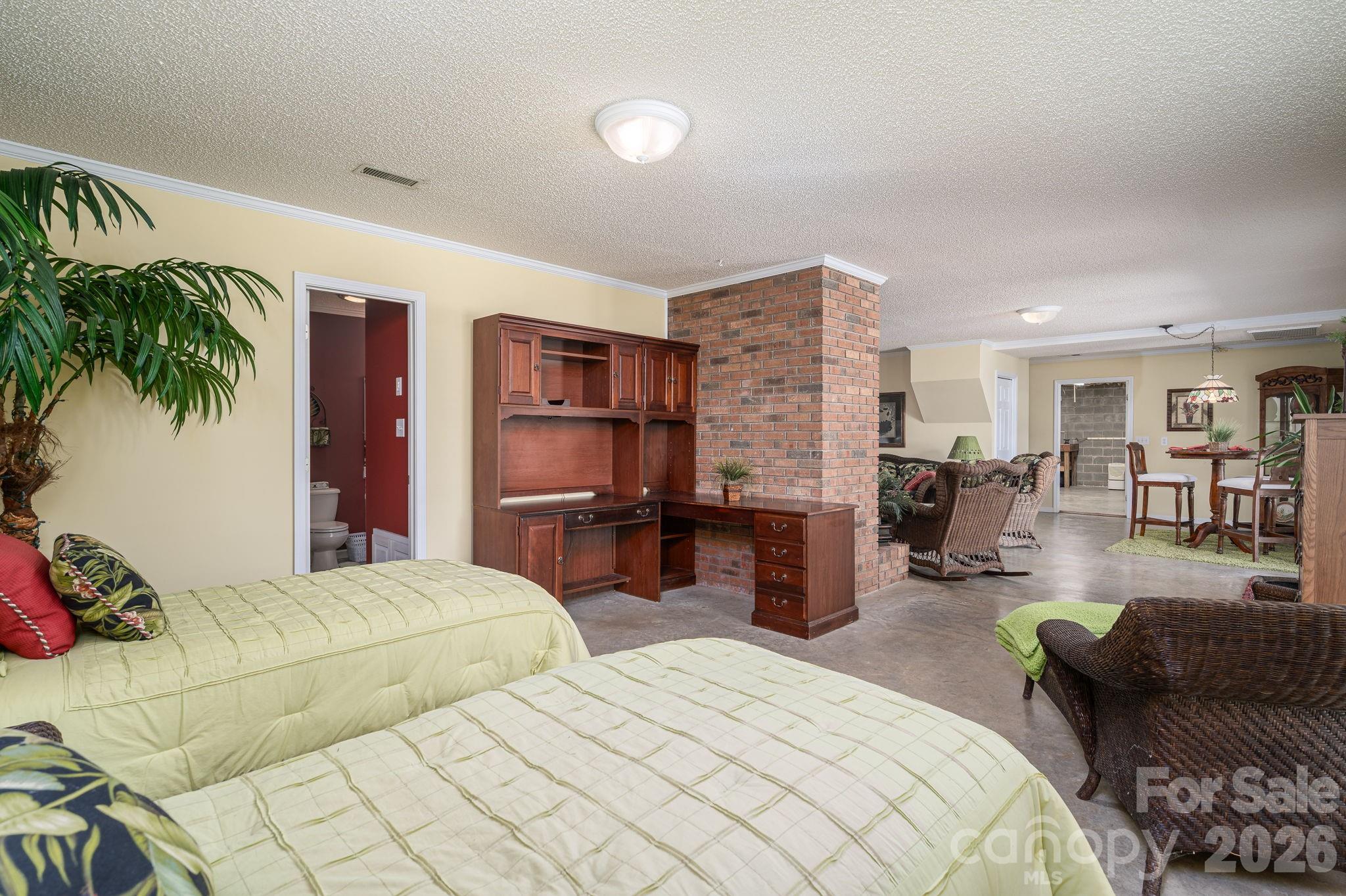 Bedroom with two beds, built-in wooden desk, brick column, and adjoining living and dining areas.